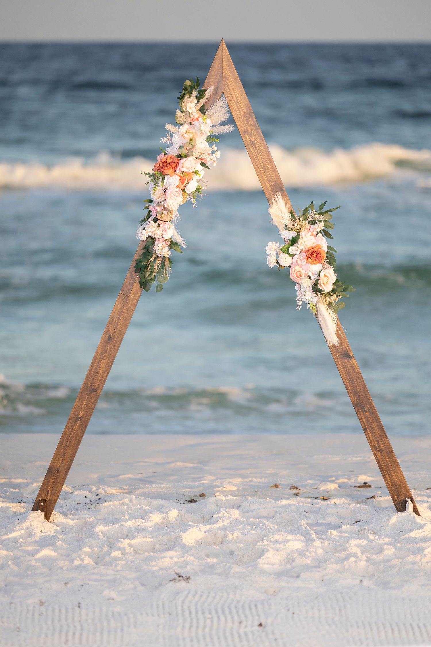 Wooden triangle wedding arch decorated with coral and white flowers stands on a sandy beach with ocean waves in background.