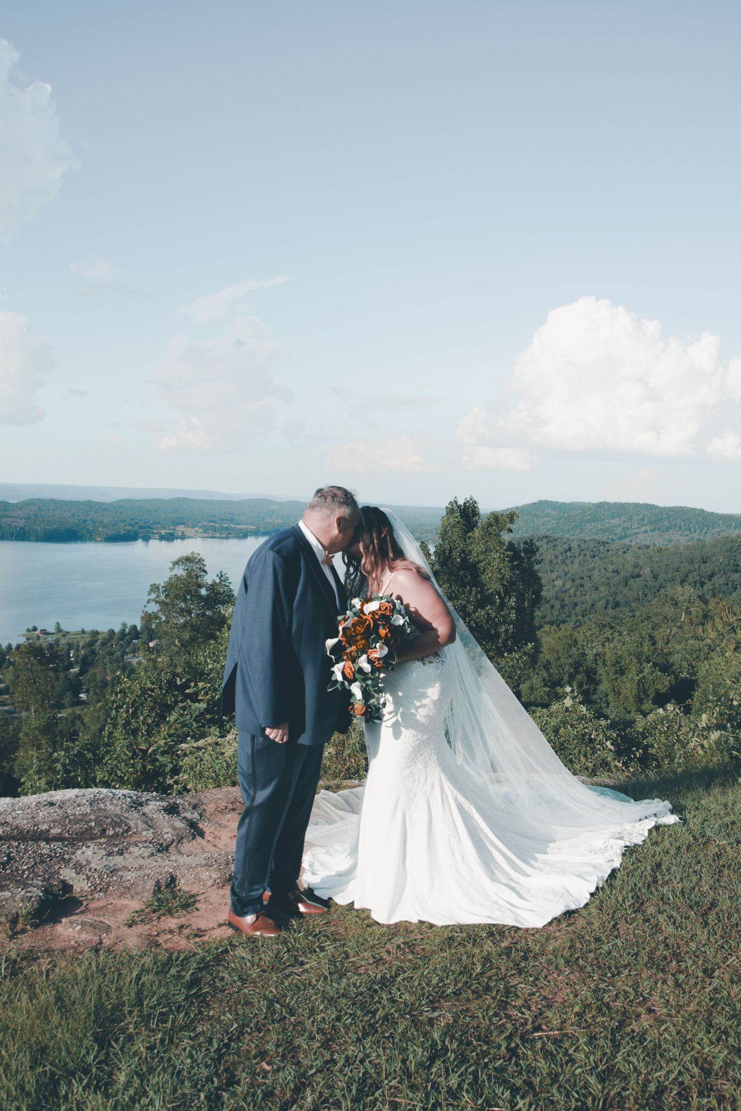 A couple in wedding attire stands on a scenic mountain overlook surrounded by lush greenery and distant hills.