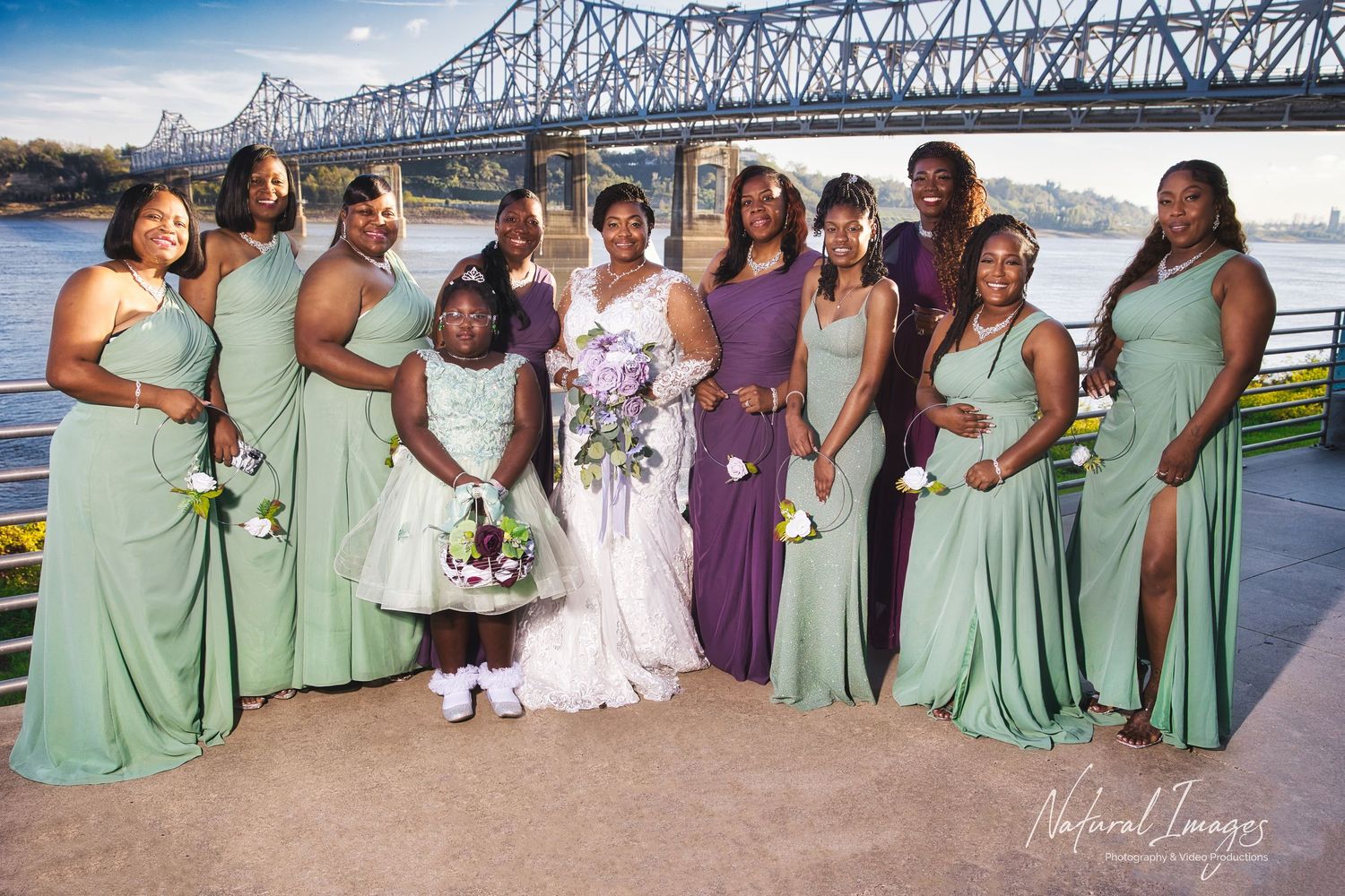 Bridal party poses outdoors near a river bridge, bride in white gown surrounded by bridesmaids in sage green and purple dresses.