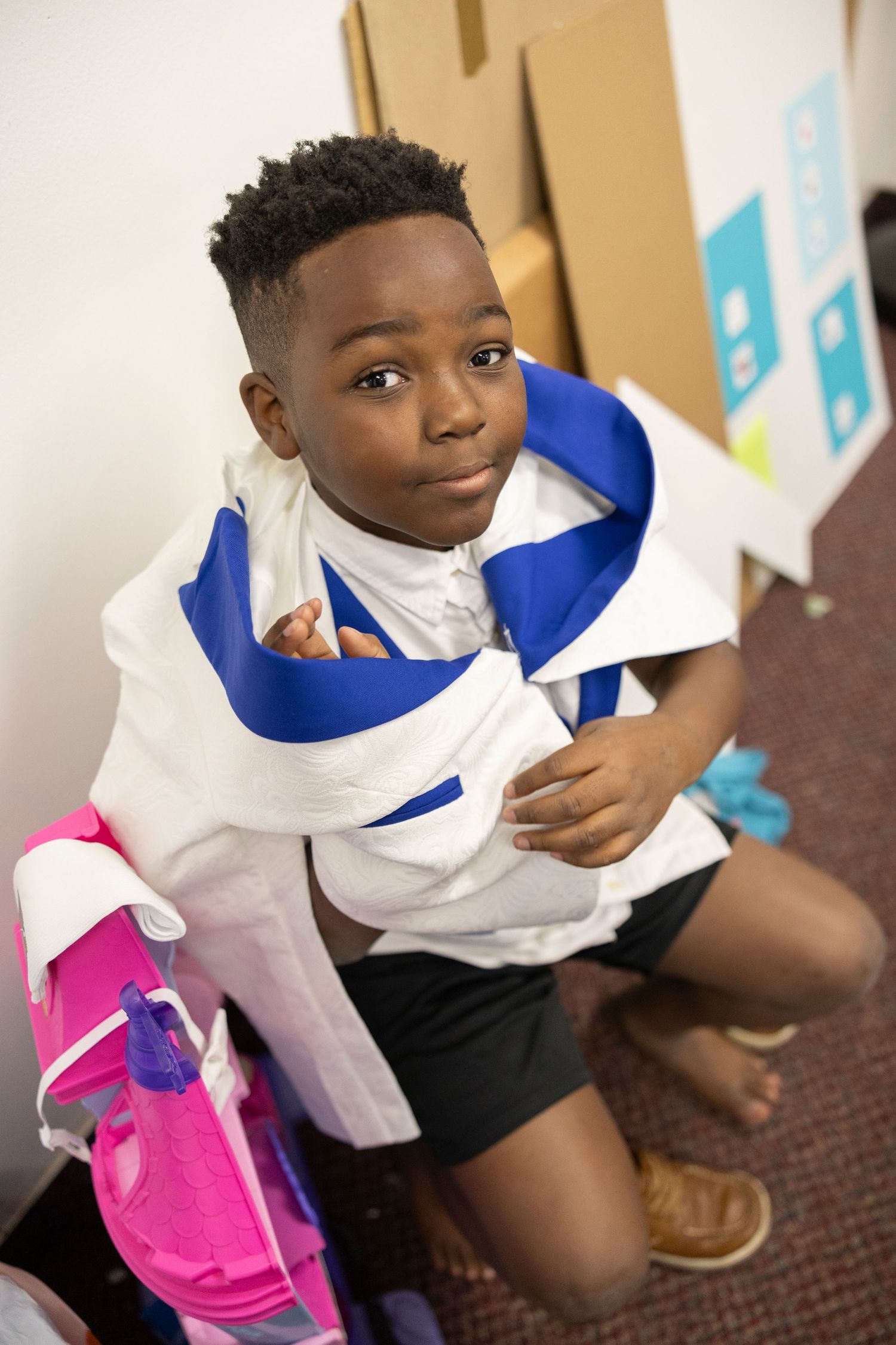 Young Black boy putting on a white and blue jacket near a pink backpack indoors.