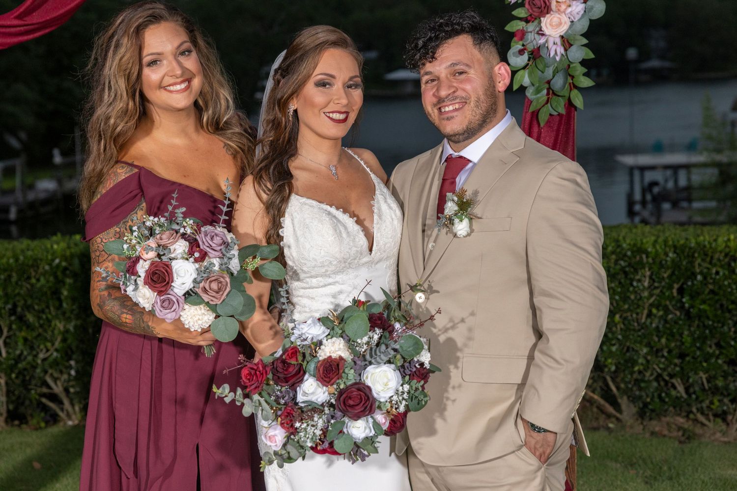Wedding party poses with burgundy and cream floral bouquets against garden backdrop at sunset.