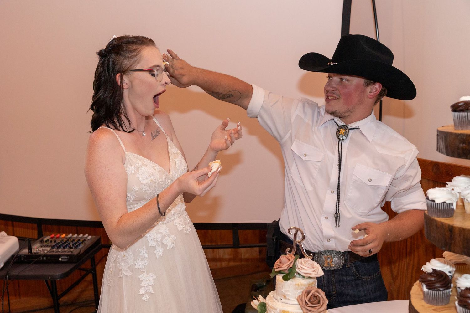 Couple shares playful moment while feeding each other wedding cake at rustic reception.