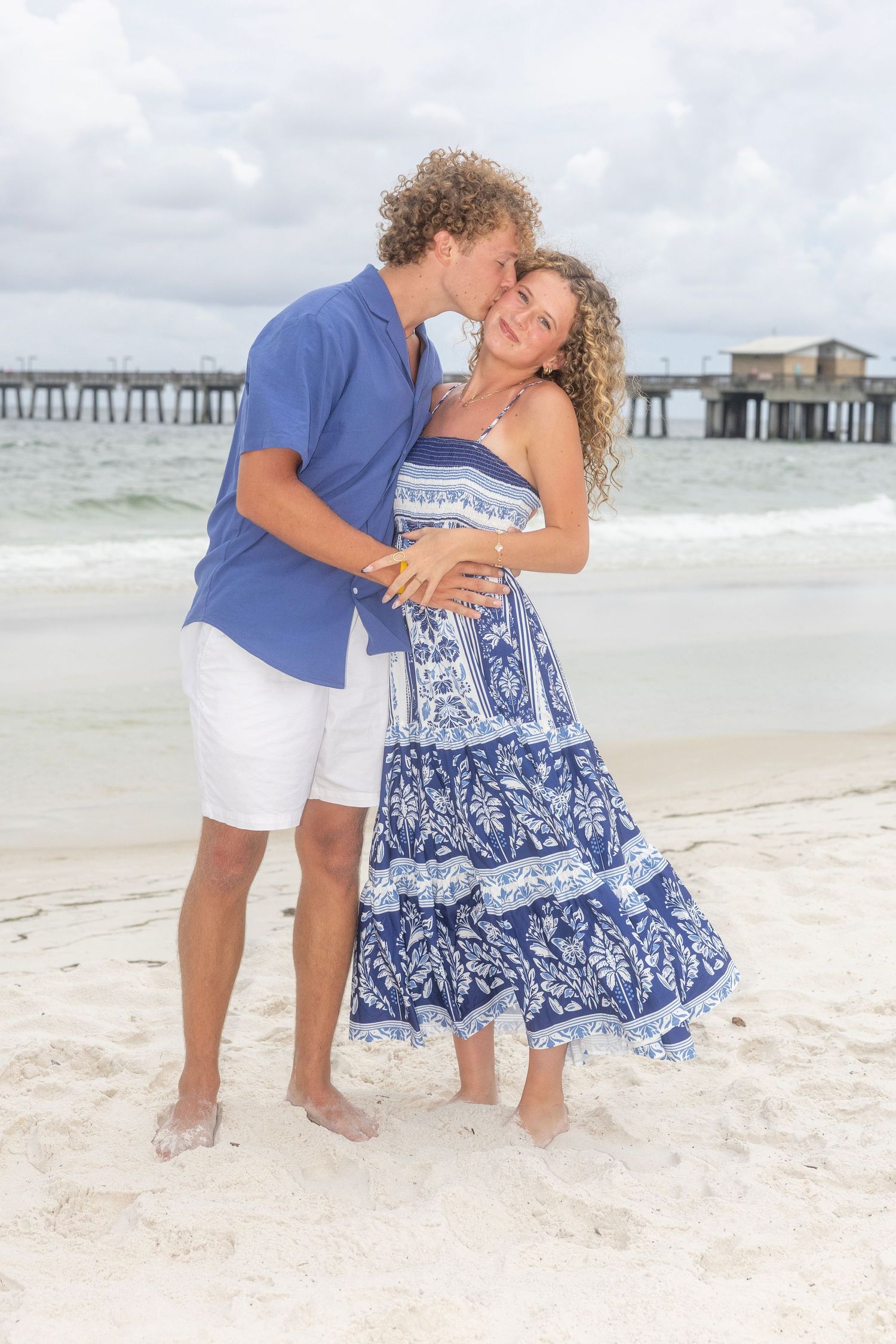Couple in blue and white outfits sharing romantic moment on beach with pier in distance.