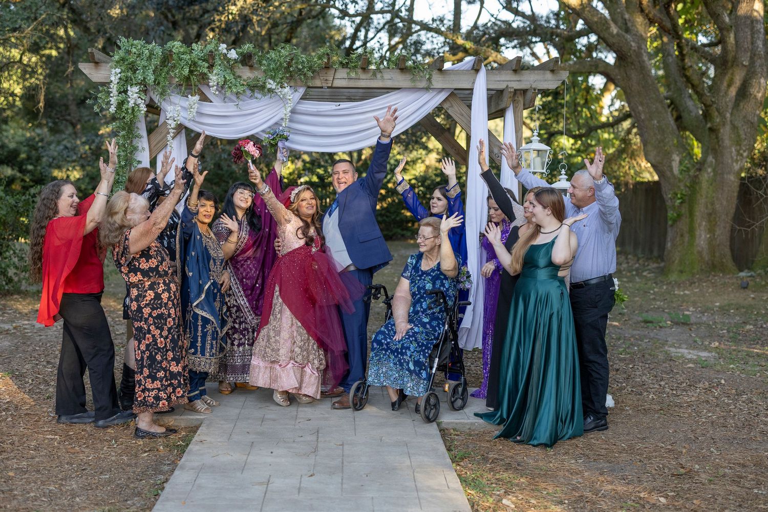 Wedding guests dance and celebrate at an outdoor reception under tall trees during a sunny autumn day.