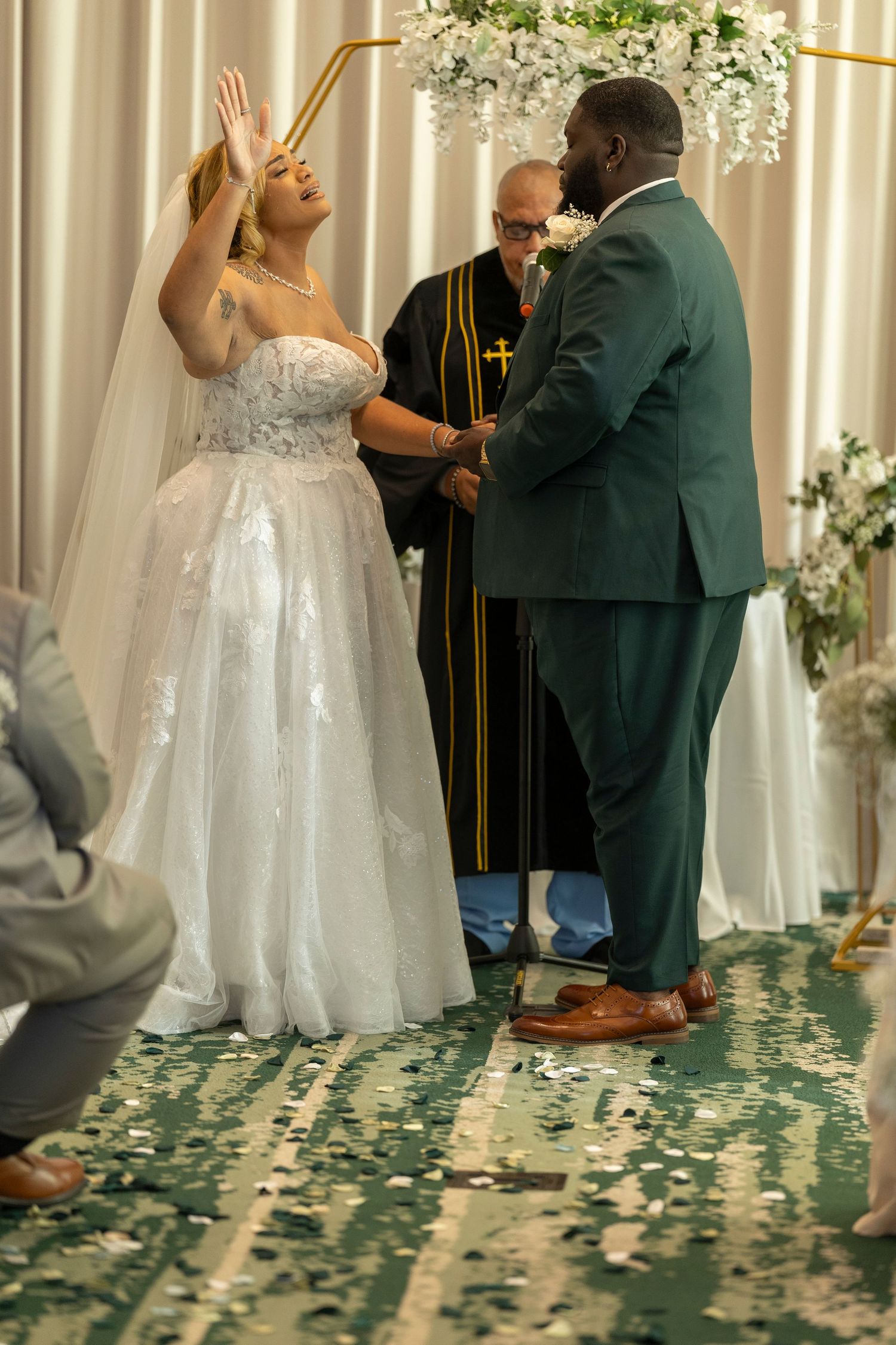 A wedding ceremony scene with a bride in white gown and groom in dark suit standing before an officiant with floral decor.