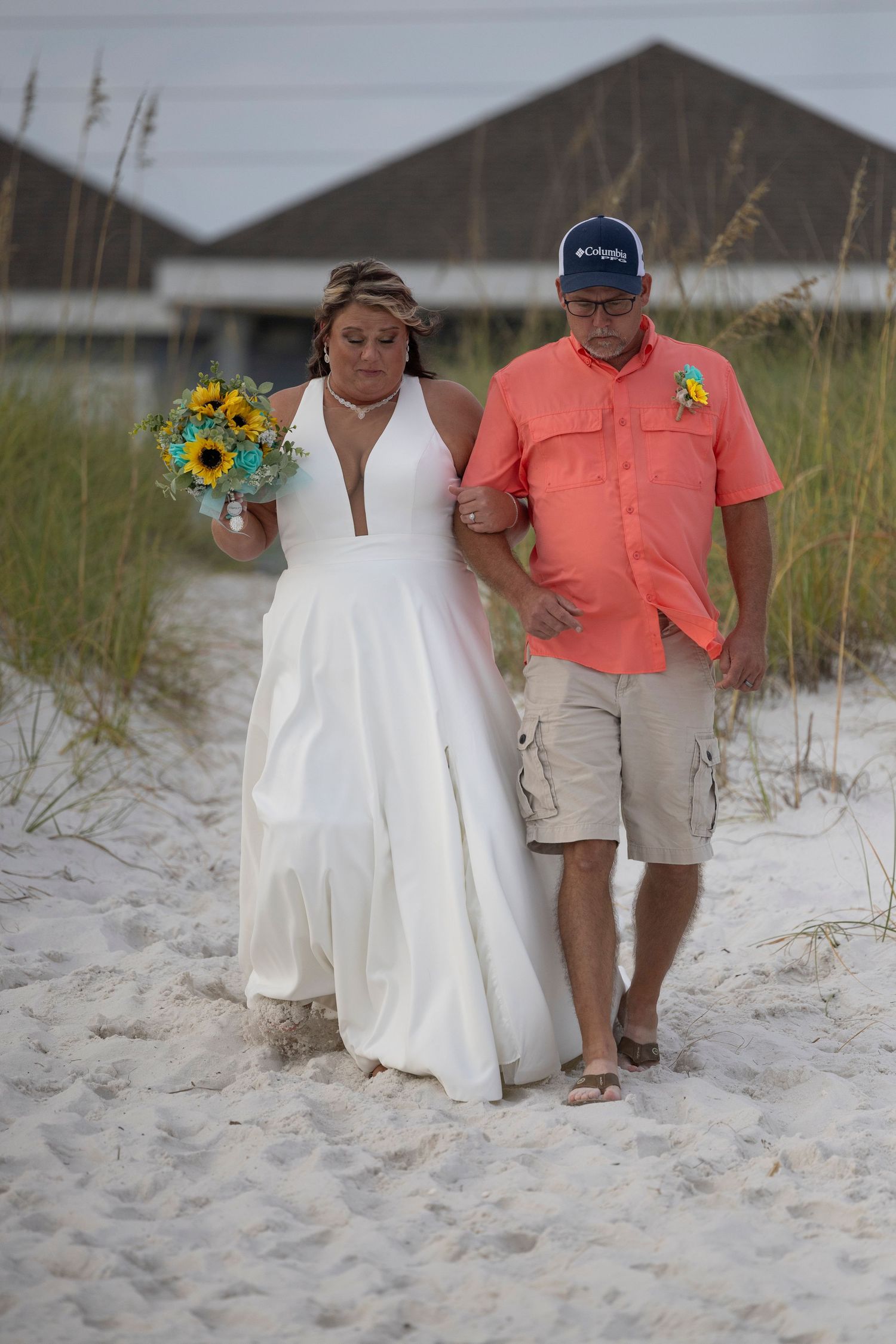 A couple walking on a beach path with sunflowers and coastal grasses, the bride in white and groom in coral.