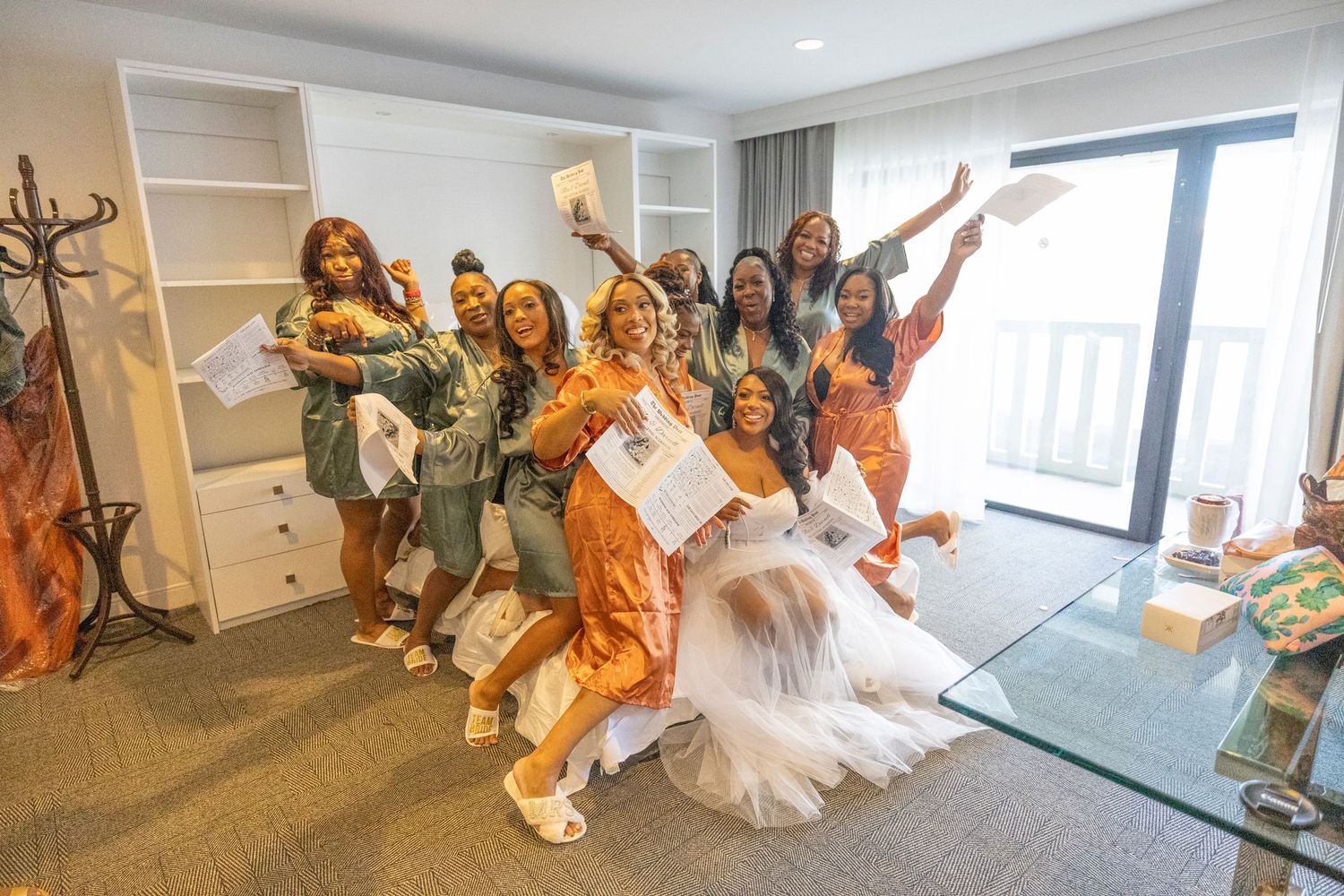 A group of people in formal attire dance together in a bright indoor space with large windows.