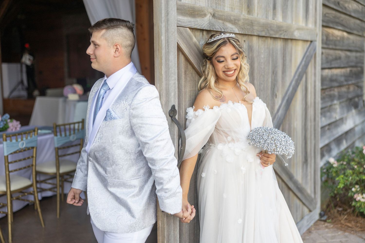 A couple in white wedding attire share a romantic moment in front of a rustic wooden barn.