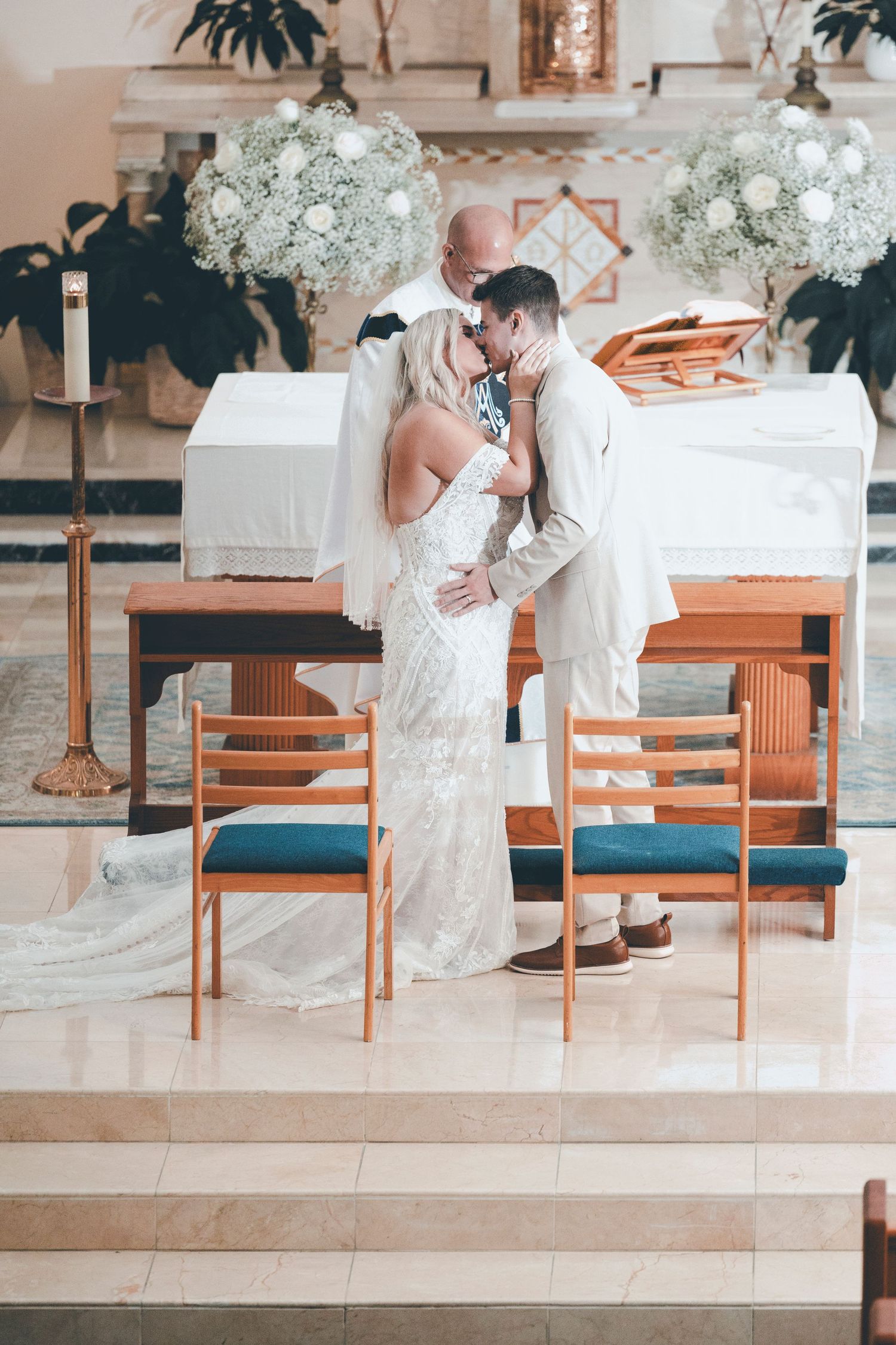 Wedding couple shares romantic moments on wooden chairs with white floral arrangements in elegant venue setting.