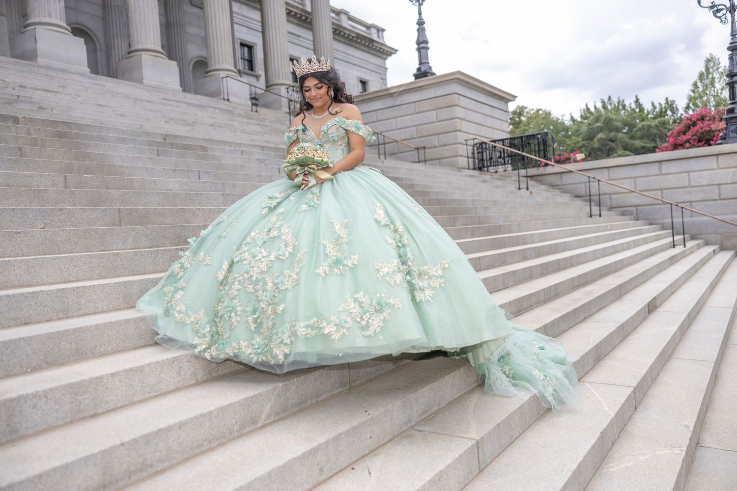 A stunning mint green quinceañera ball gown with gold embroidery sparkles on grand stone steps of a building.