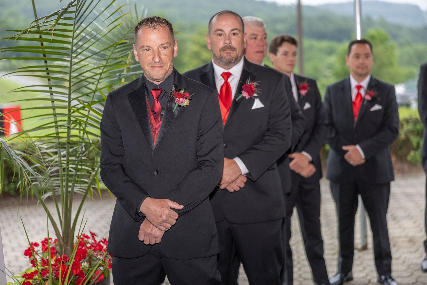 A wedding party in formal black suits and red accents poses outdoors against a scenic mountain backdrop.