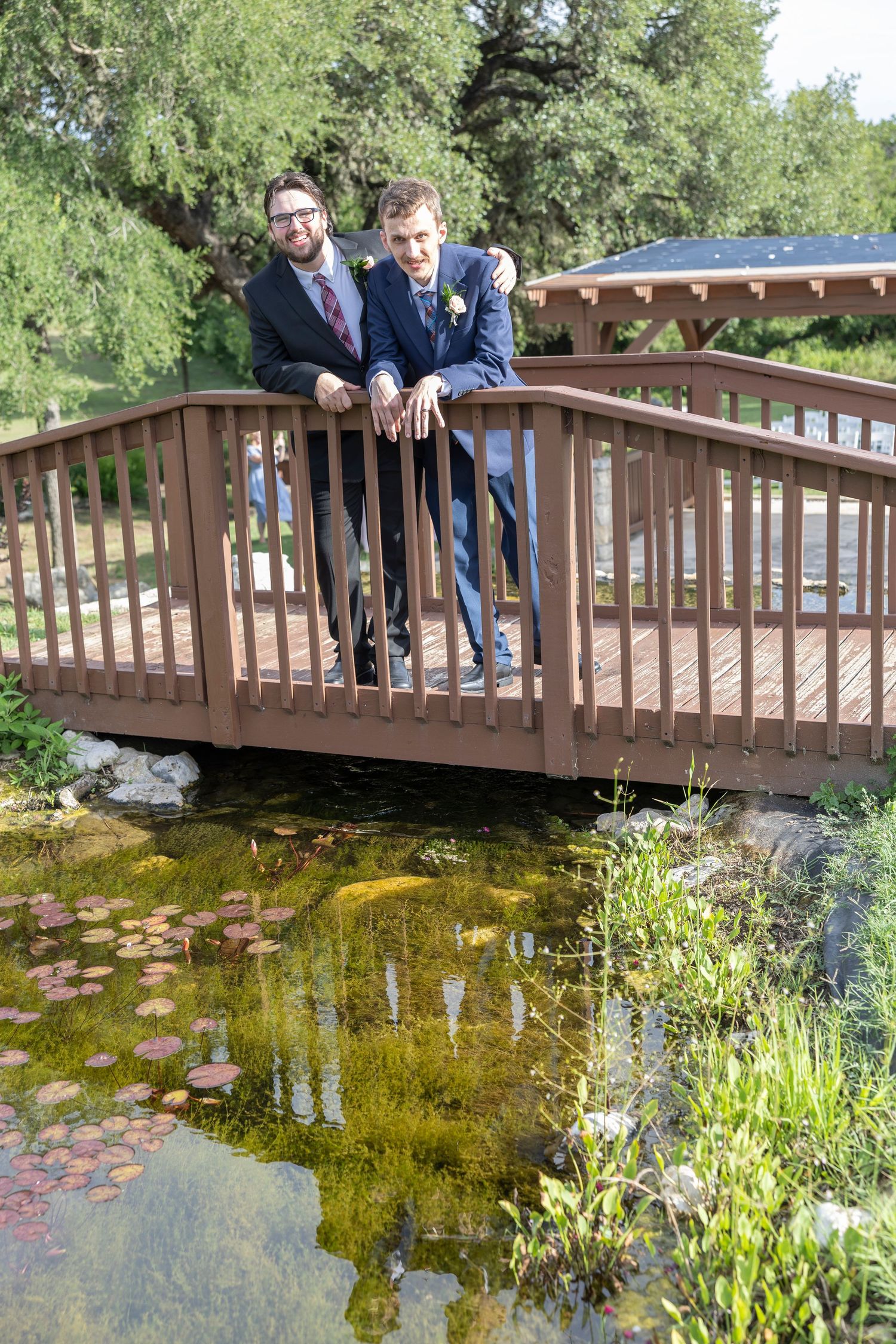 People lean over a wooden bridge railing to look at a pond with green vegetation and reflective water below.