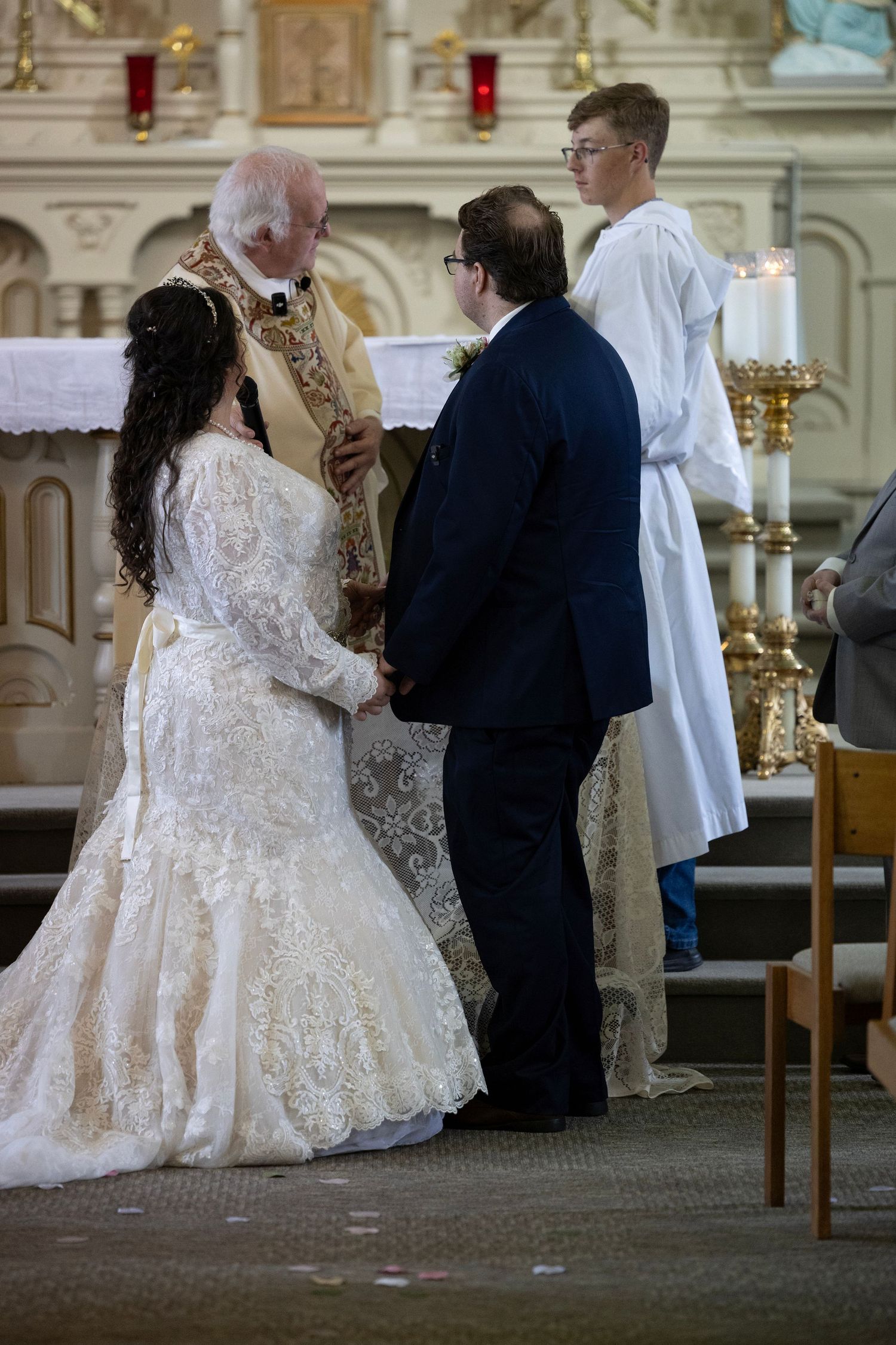 A couple in formal wedding attire exchanges vows during a traditional church ceremony with candlelit altar.