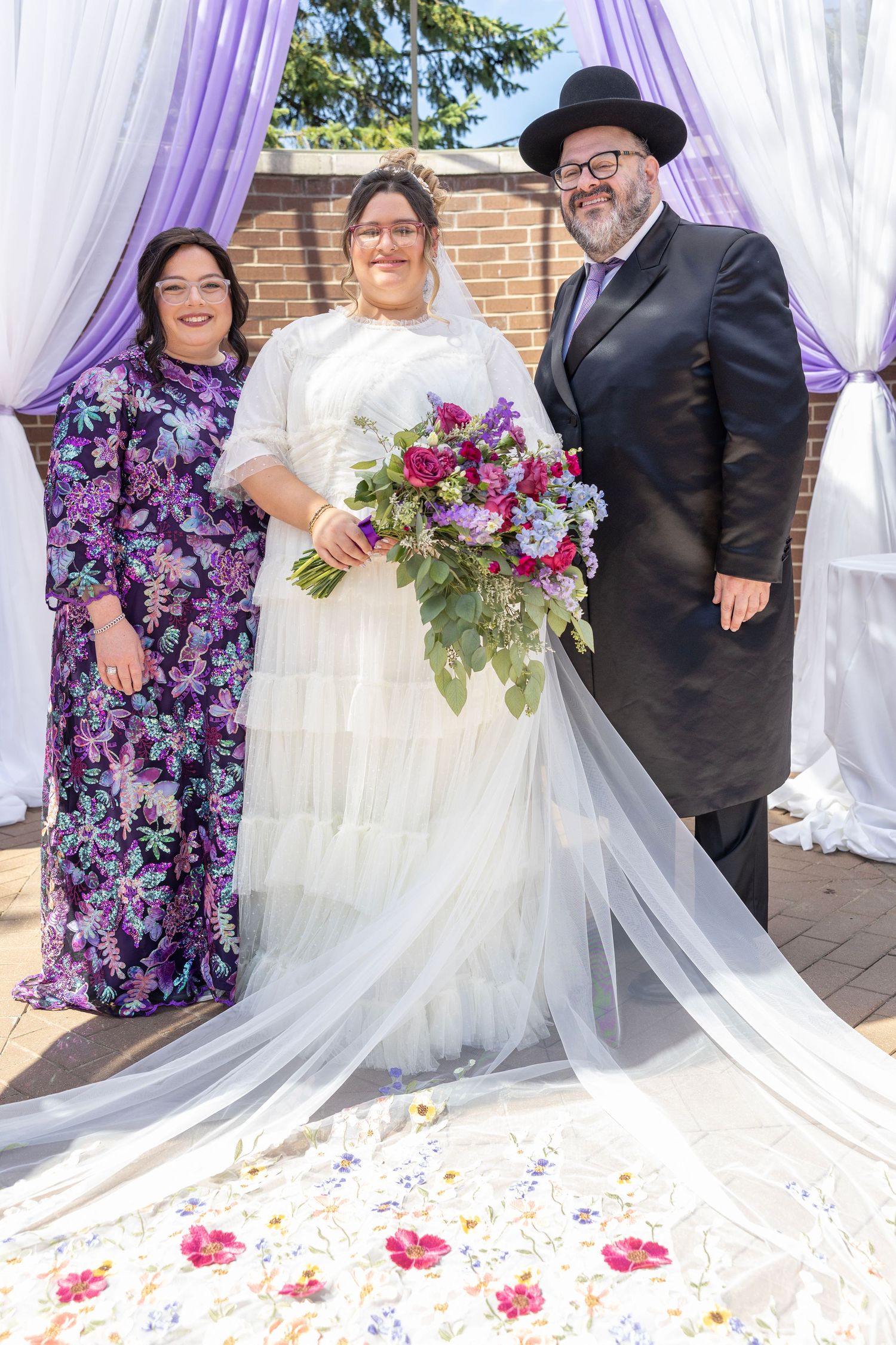 Wedding party poses together under purple and white draped fabric with floral bouquets and scattered rose petals.