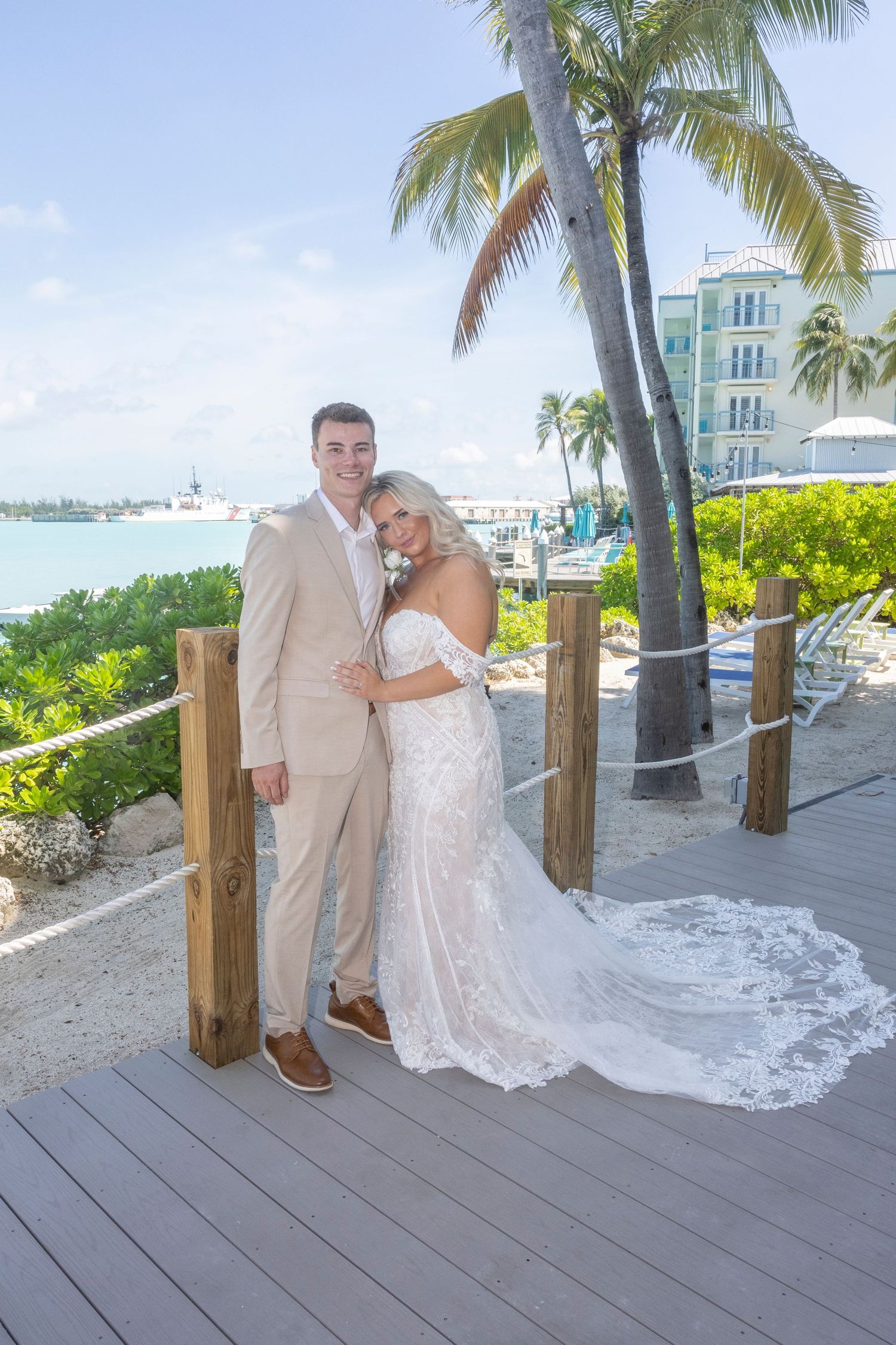 A couple poses on a wooden boardwalk with palm trees and beach scenery in the background.