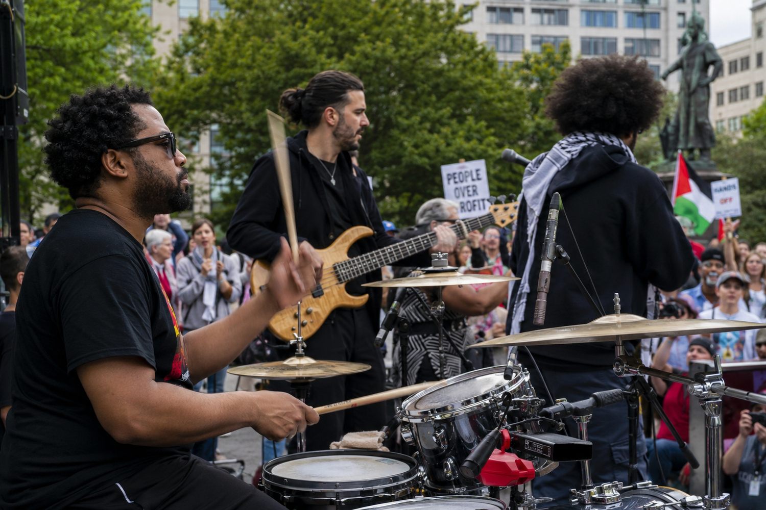 Musicians perform on drums and guitar at an outdoor pro-Palestine rally with crowds and flags in the background.