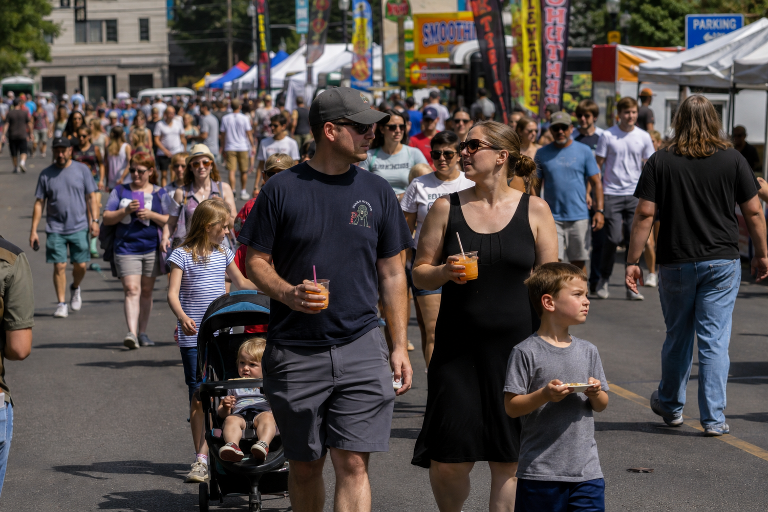 A family with a stroller and young child enjoys food and drinks while walking through a sunny outdoor street fair.