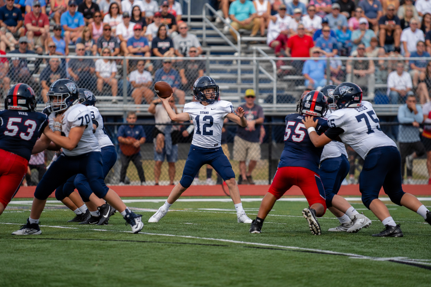 High school quarterback number 12 throws a pass while offensive linemen block during a football game.