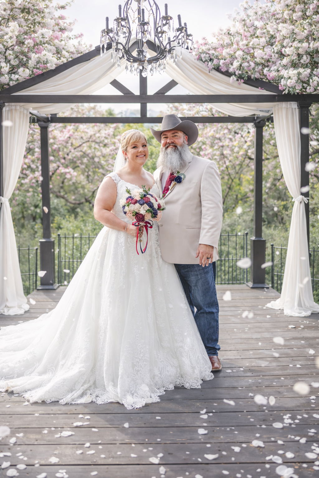 Bride and groom pose under floral gazebo with chandelier, she in white gown with bouquet, he in cowboy hat and jeans.