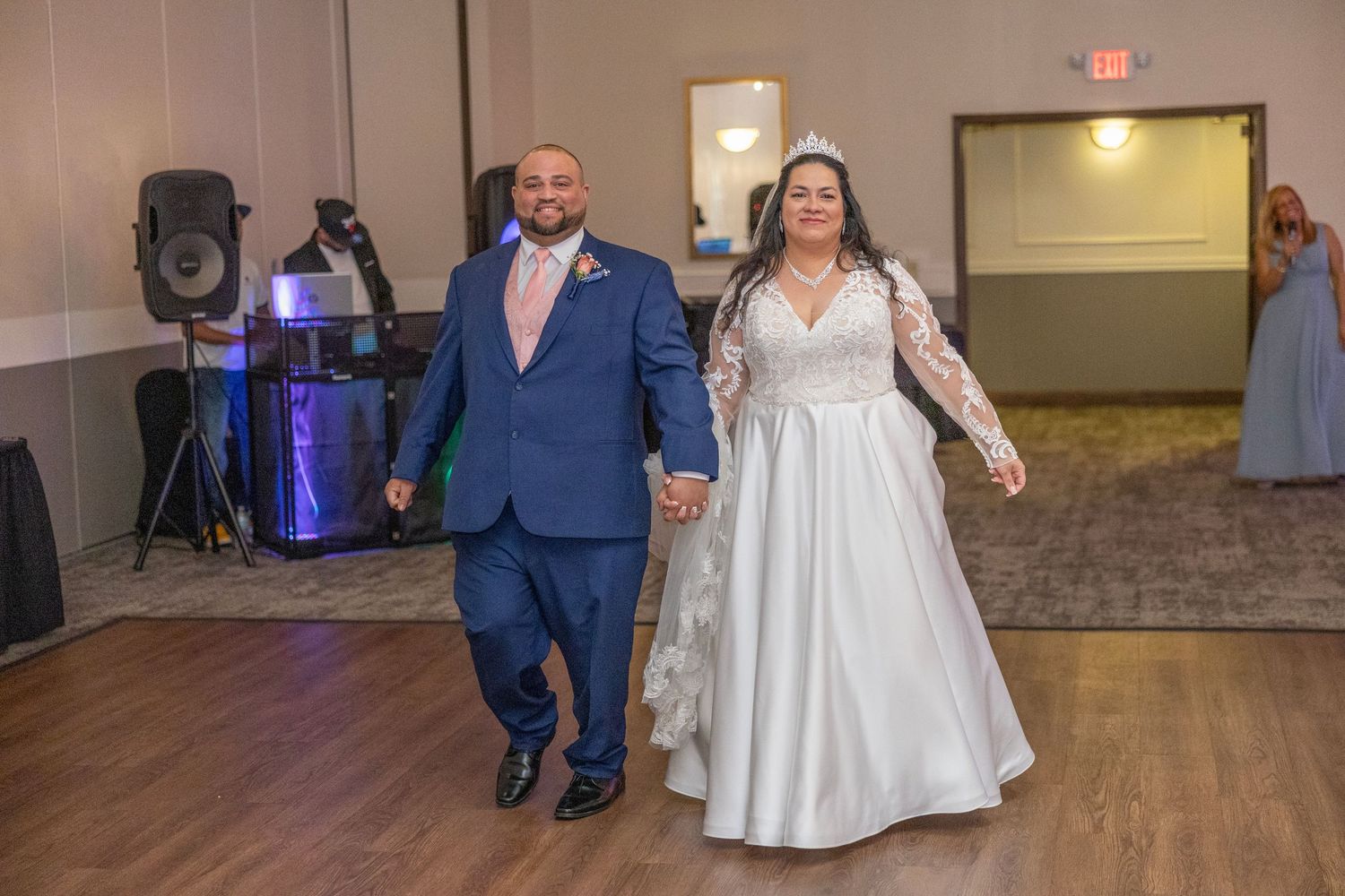 Bride and groom make grand entrance at wedding reception, bride in white gown, groom in blue suit with pink tie.