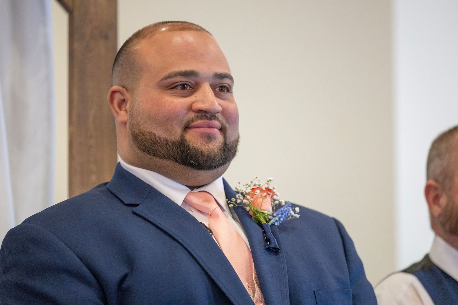 A smiling groom in a navy blue suit with a peach tie and floral boutonniere stands at the altar on his wedding day.