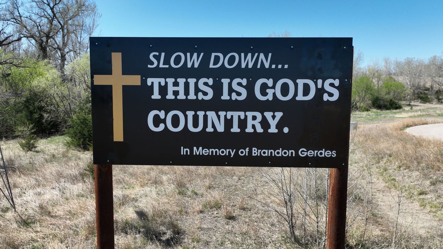 Roadside memorial sign reading Slow Down This Is Gods Country in memory of Brandon Gerdes with a cross symbol.