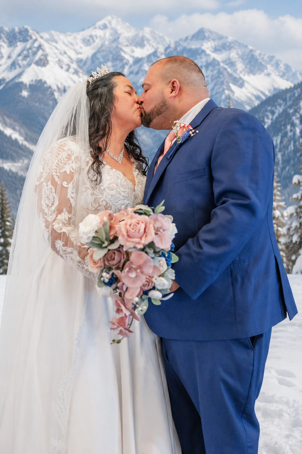 Bride and groom share first kiss with snow-capped mountains backdrop, bride in lace gown holding pink bouquet, groom in blue suit.