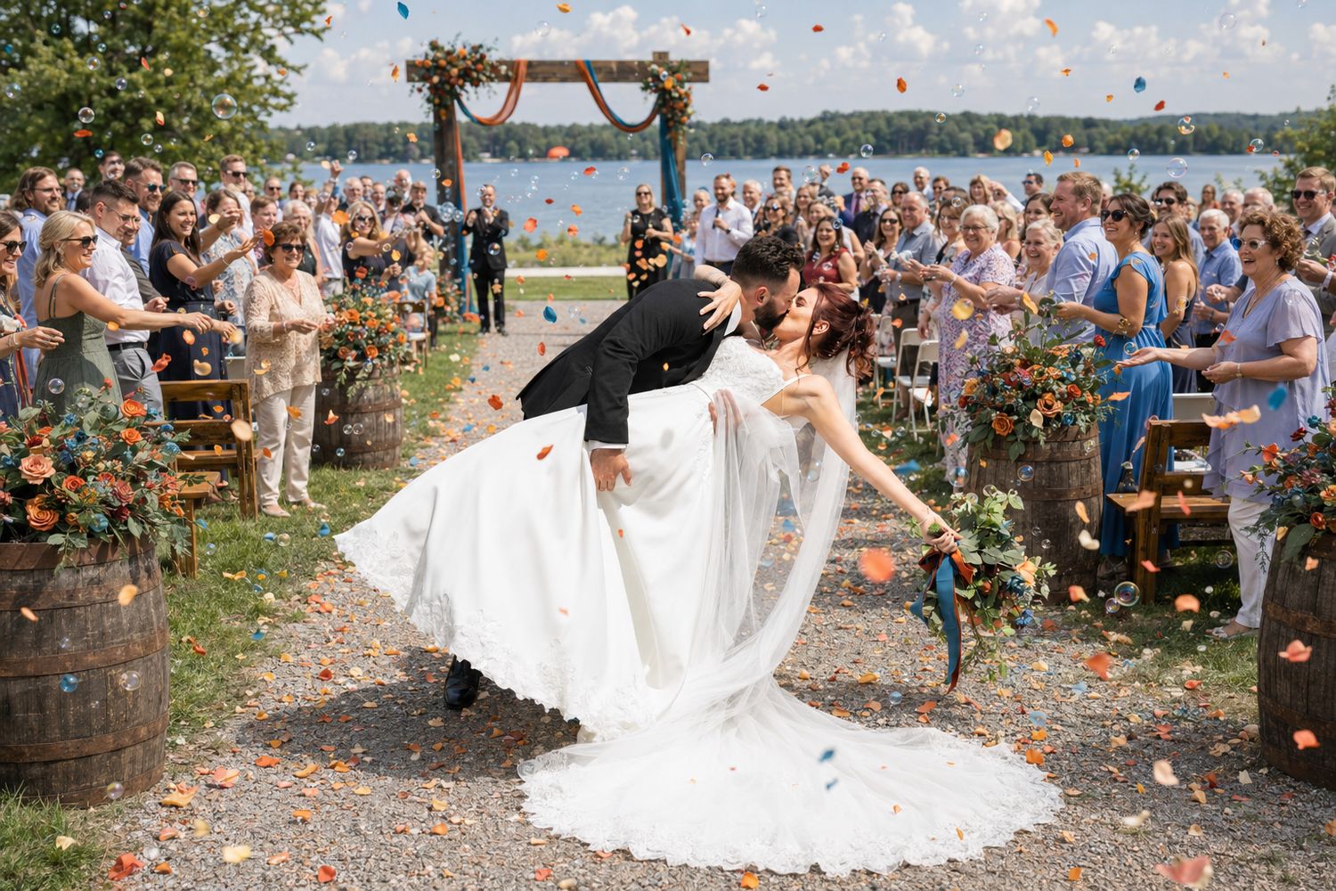 Groom dips bride during joyful wedding recessional as guests toss colorful flower petals outdoors.