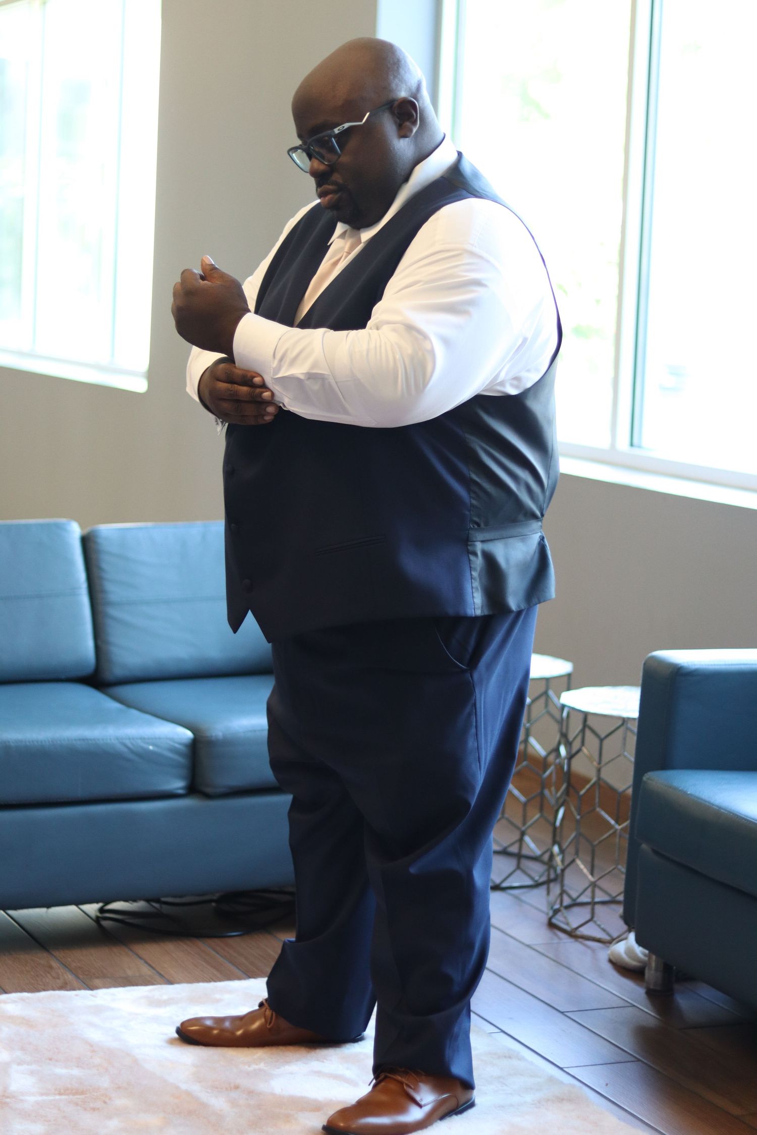 A well-dressed man in a navy blue vest and trousers adjusting his cufflinks in a modern lobby.