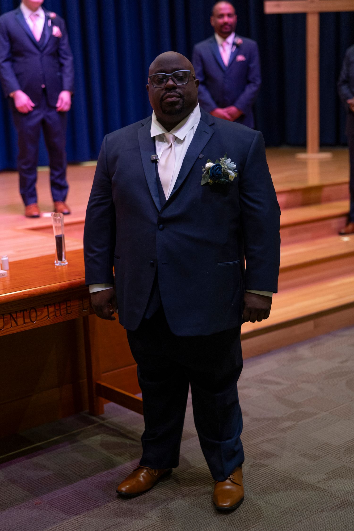 Man in navy blue suit with boutonniere and tan tie stands at wedding ceremony in church.