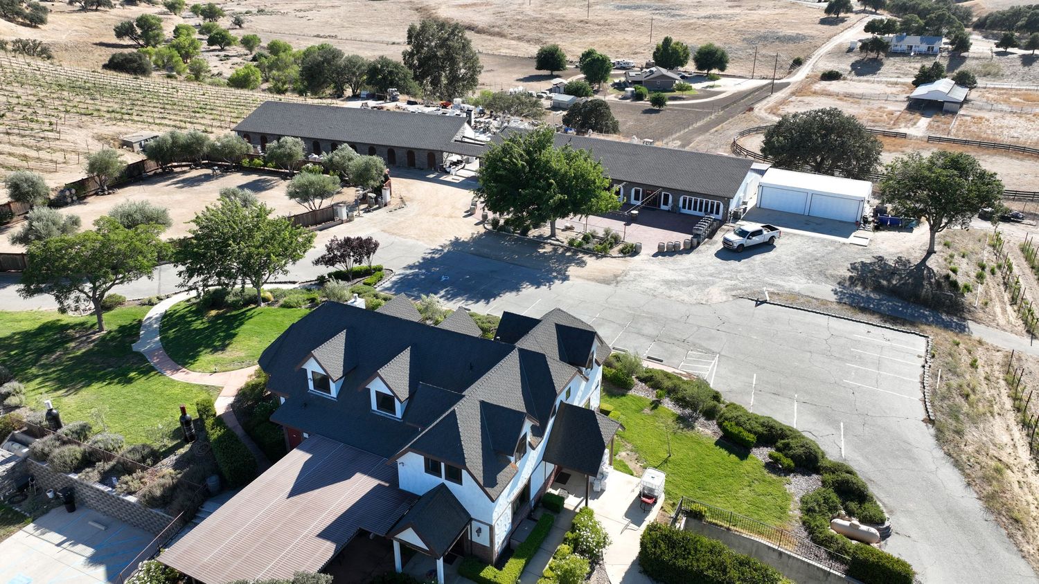 Aerial view of a rural ranch property featuring a large home, outbuildings, paved driveway, and sparse trees in a dry landscape.