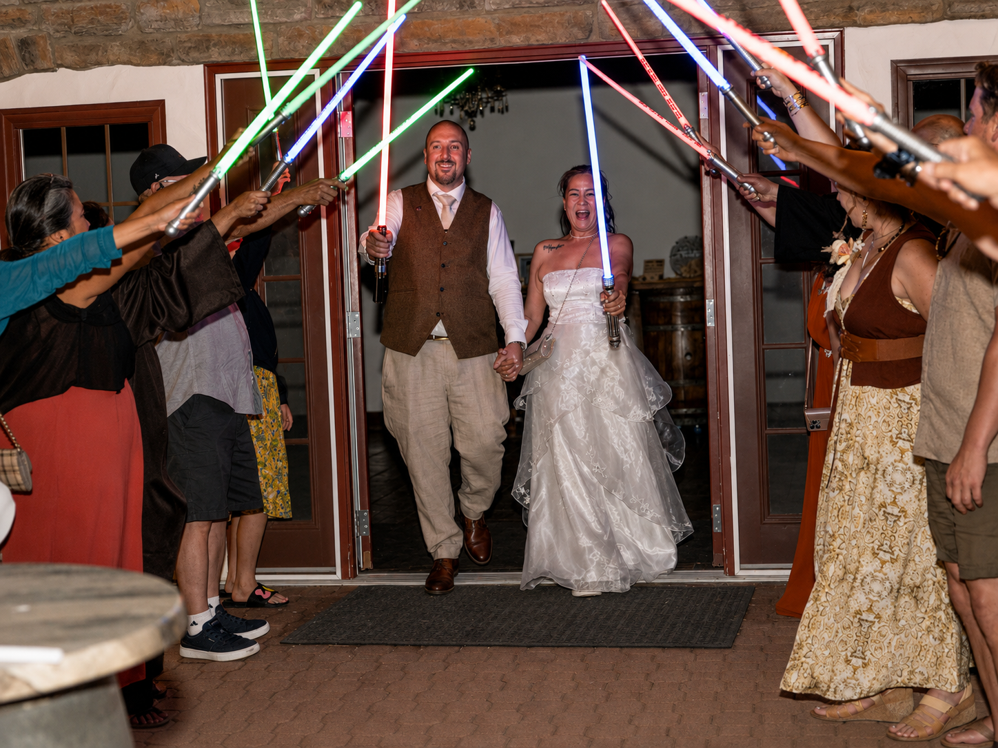 Newlyweds exit wedding venue under colorful lightsaber arch held by guests in Star Wars themed wedding celebration.