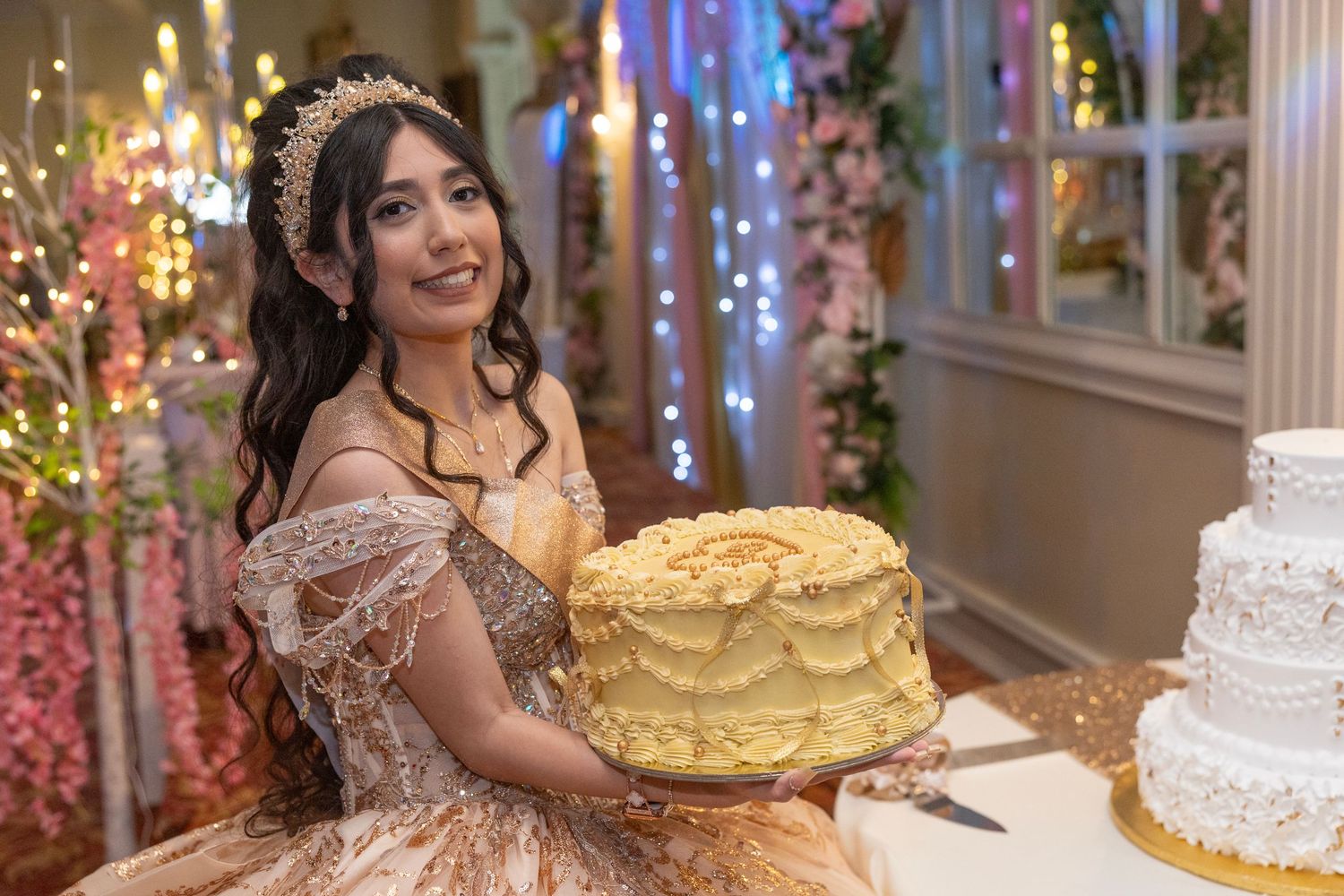 Young woman in golden quinceañera gown holding yellow cake at elegant decorated celebration party.