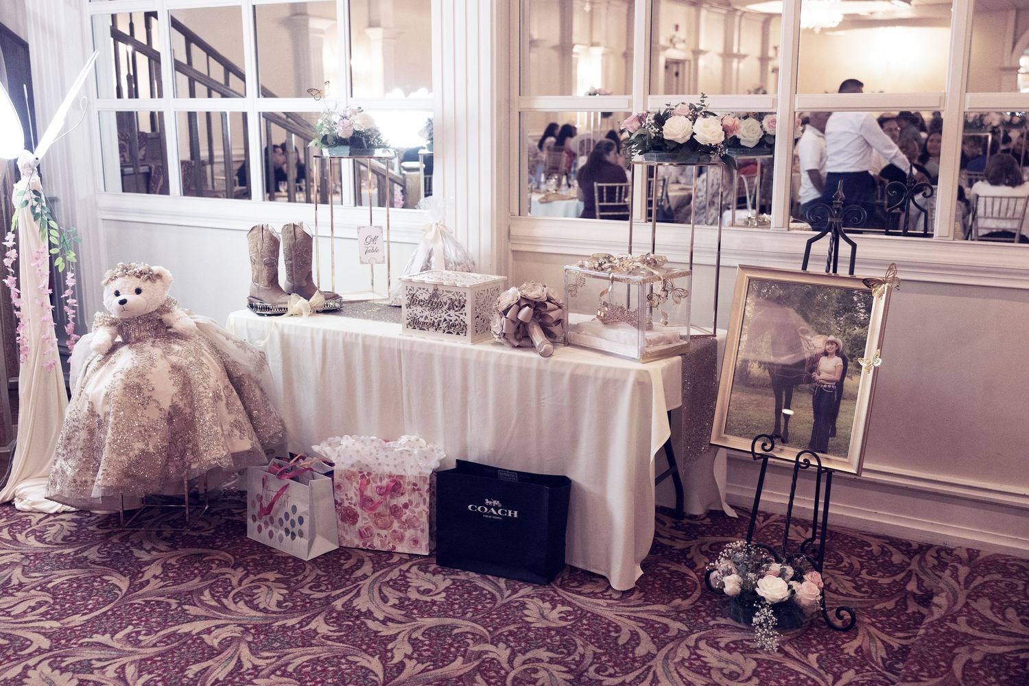 Elegant wedding display table with lace decorations, cowboy boots, teddy bear, and framed photo in a ballroom.