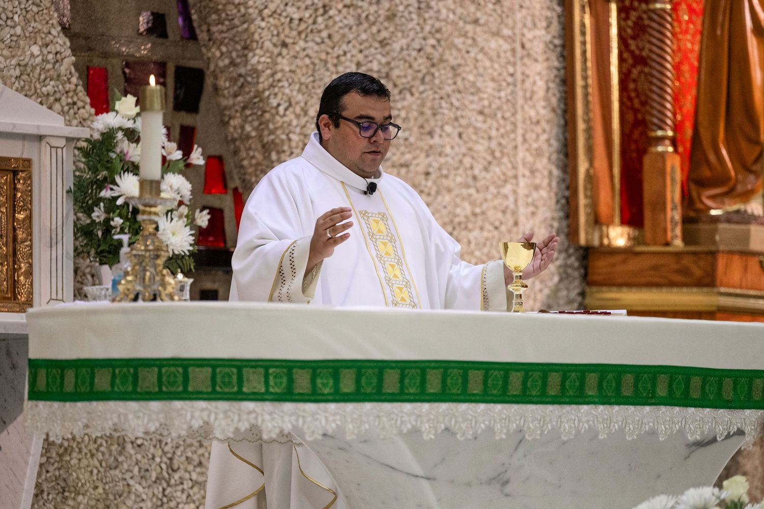 Catholic priest in white vestments delivering a sermon at an ornate altar during a religious Mass ceremony.