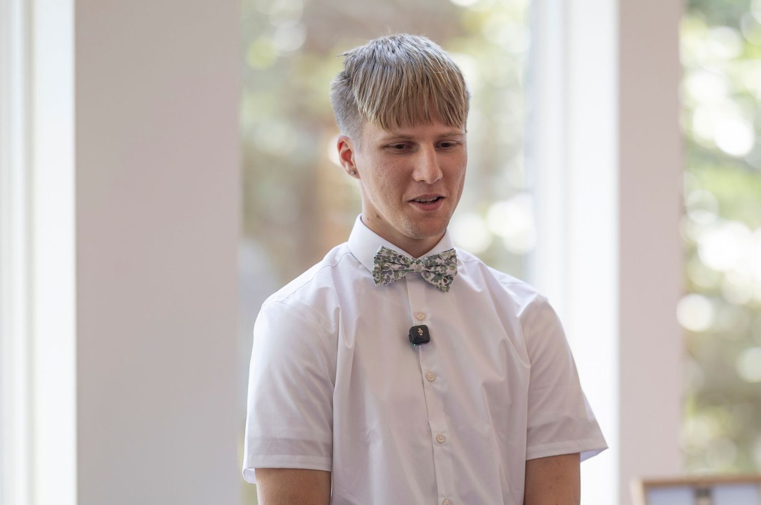 Young man with blonde hair wearing a white short-sleeve shirt and patterned bow tie with a lapel microphone.