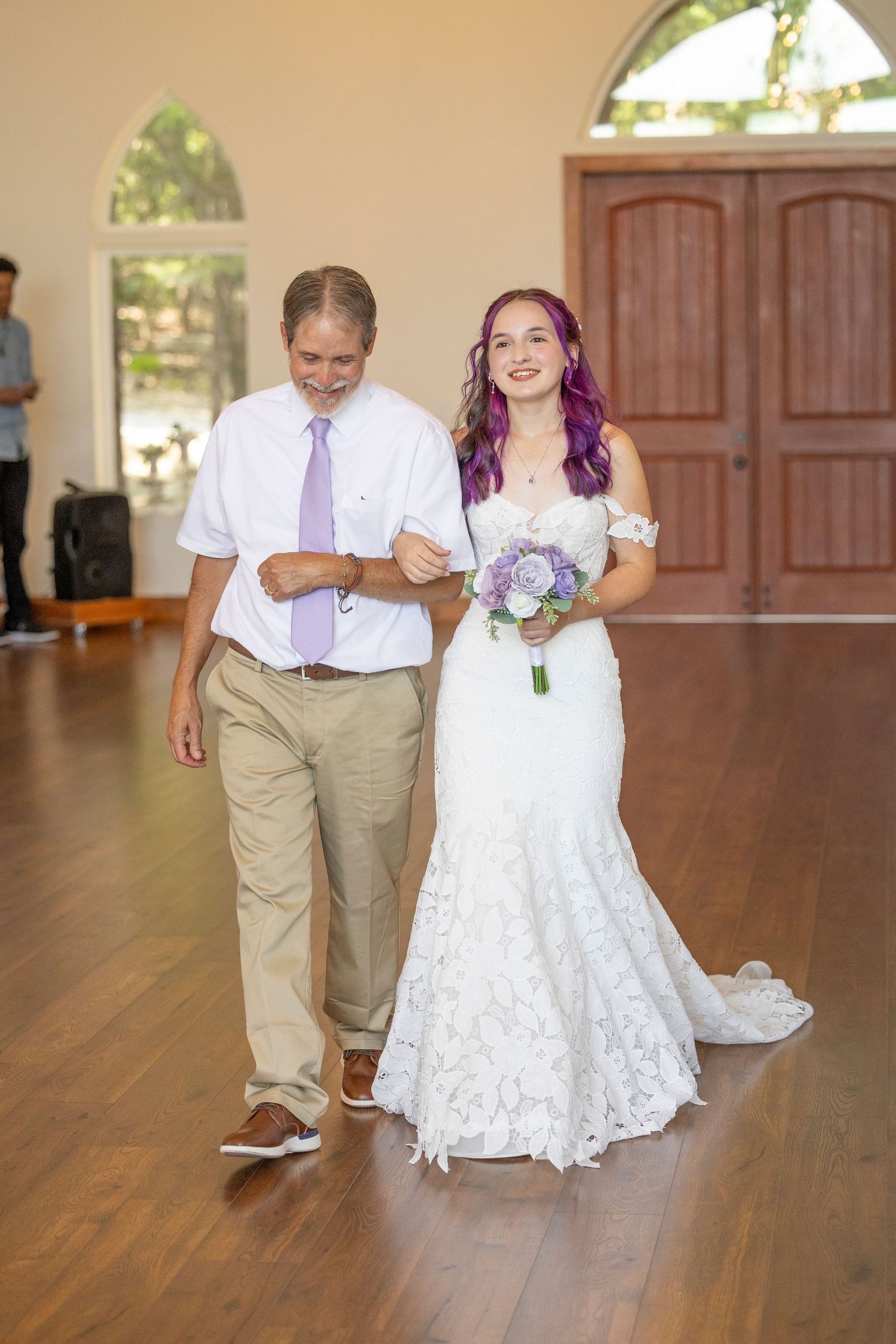 Bride with purple hair in white lace gown holding bouquet walks down aisle with father at wedding ceremony.