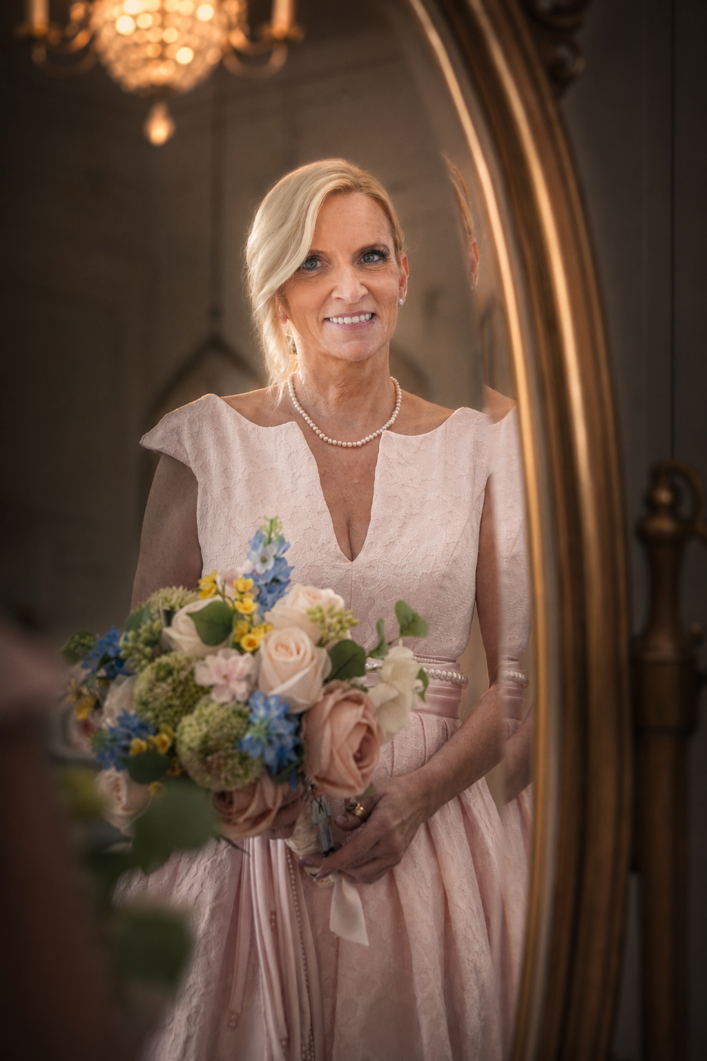 Elegant bride in blush gown holding colorful bouquet, smiling in ornate gold mirror reflection.