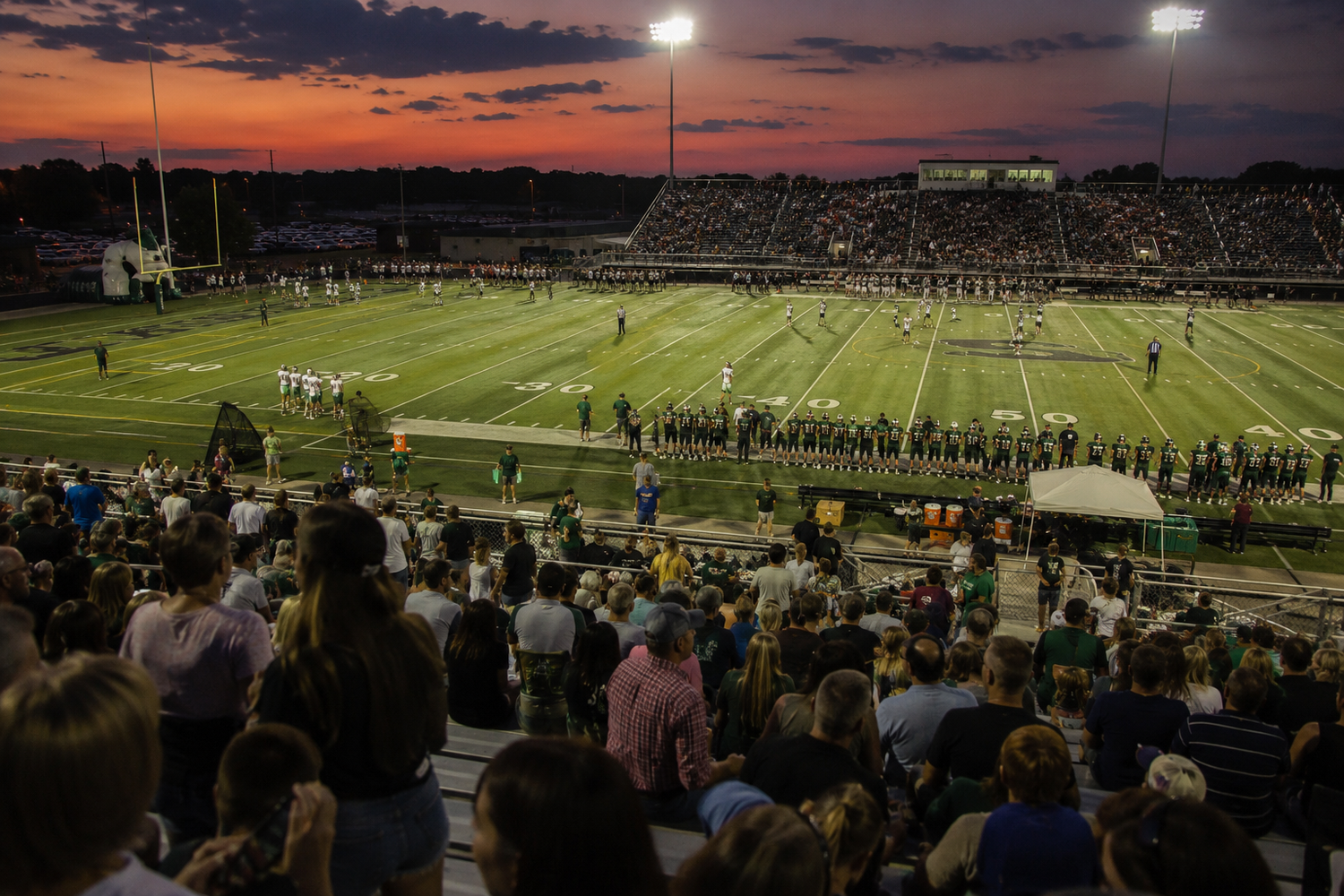 Aerial view of a packed football stadium at sunset with marching band performing on the field.