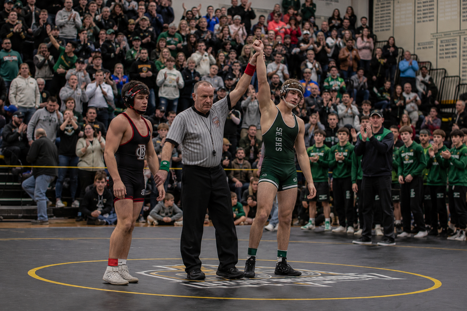 Referee raises the hand of a victorious wrestler in green uniform after a high school wrestling match.