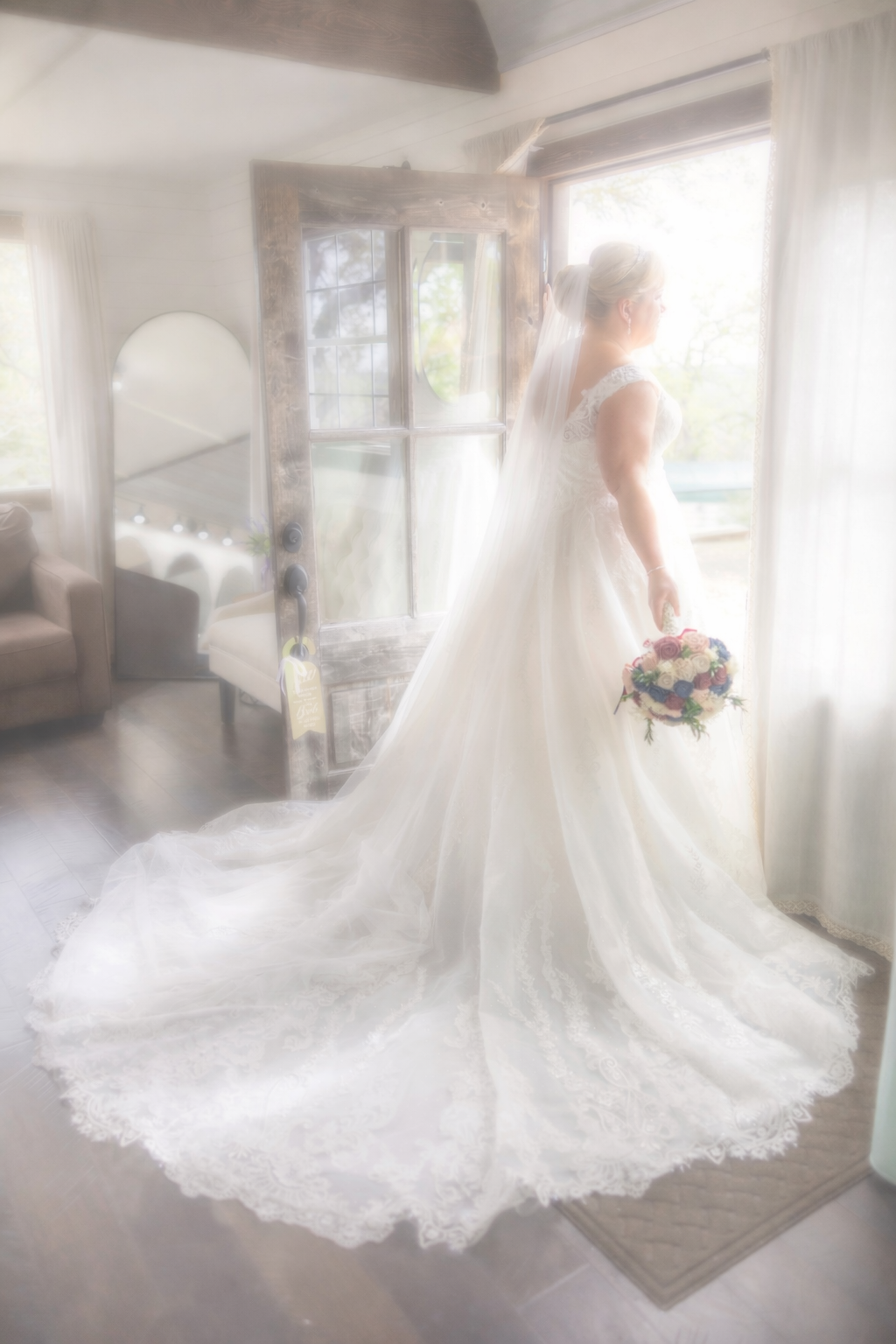 Bride in elegant white lace wedding gown with long train holds bouquet while standing by bright window.