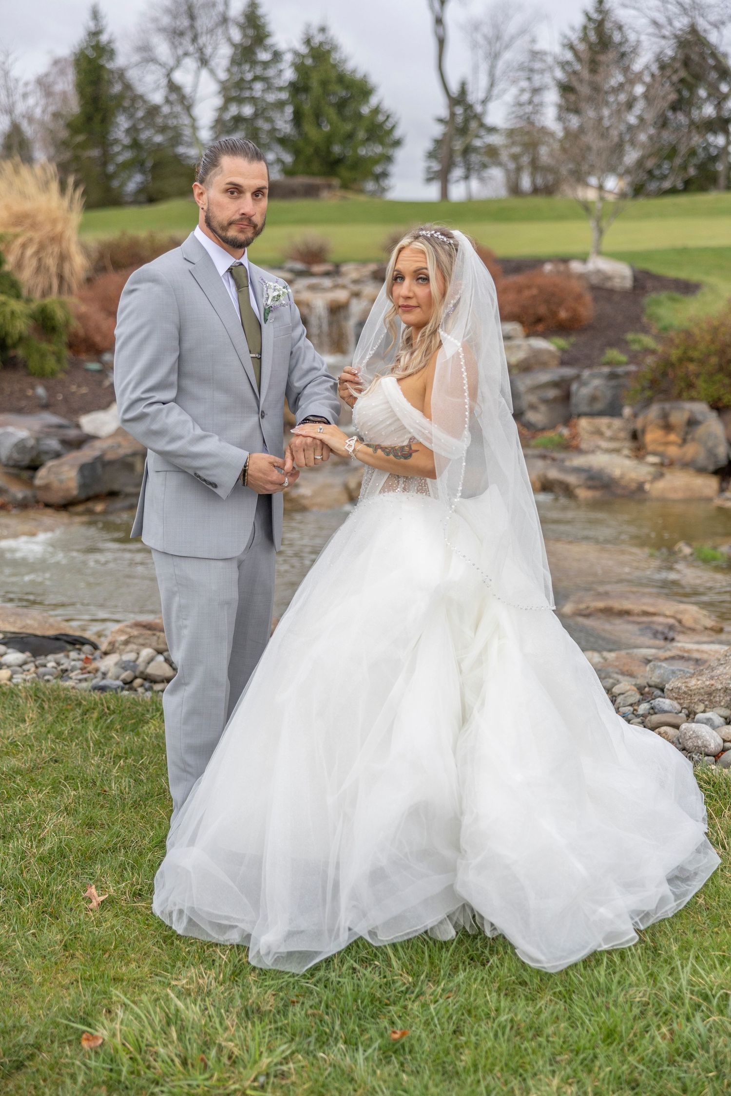 A bride in a flowing white gown and groom in a light gray suit pose together on grassy grounds with a rock garden backdrop.