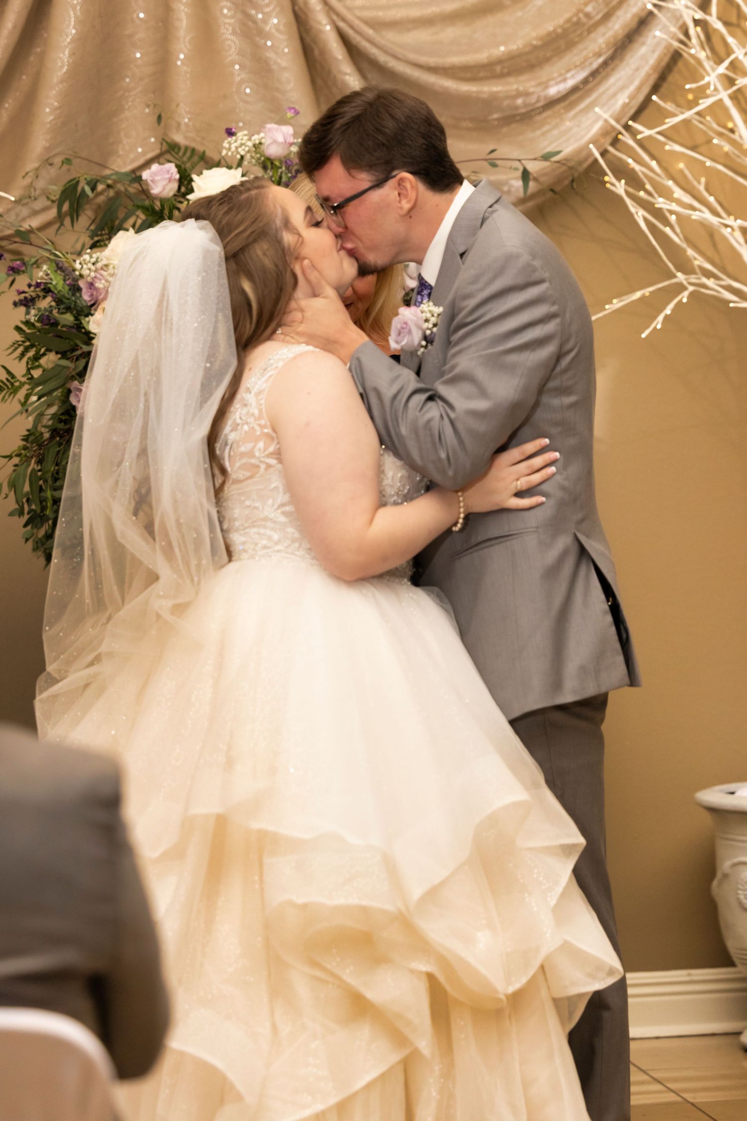 A romantic wedding kiss between newlyweds, with the bride wearing a lace gown and the groom in a grey suit.