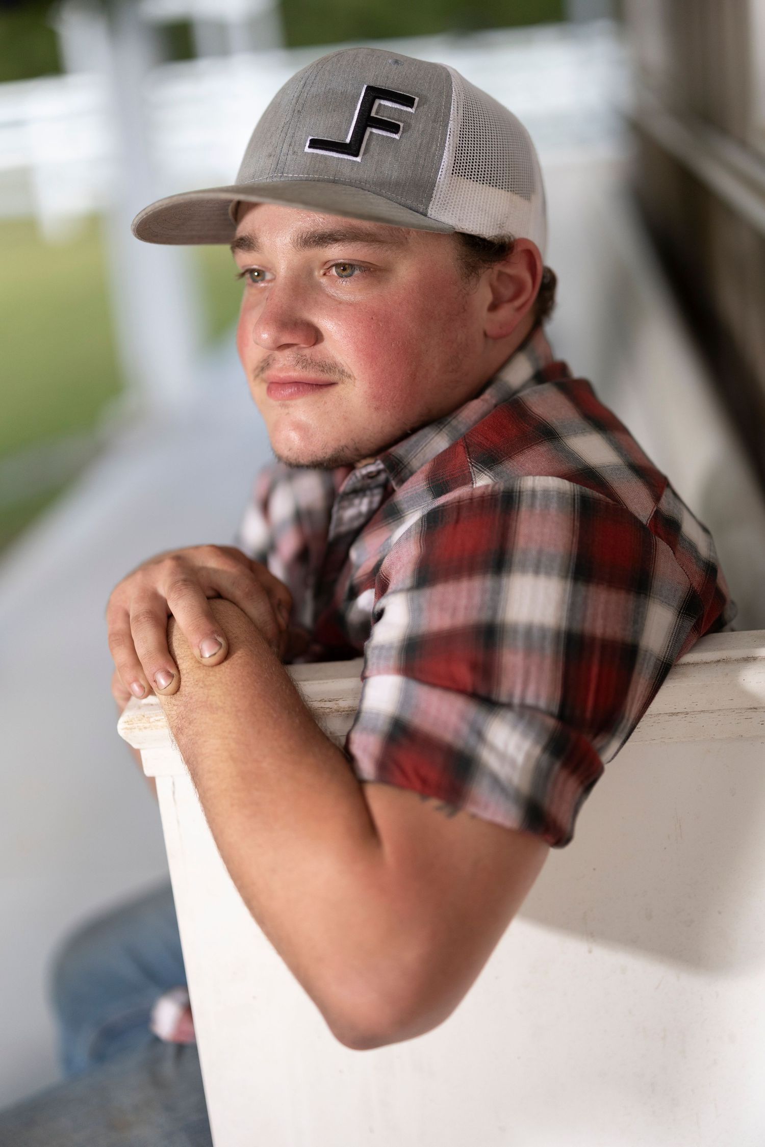 A young person in a plaid shirt and mesh baseball cap with letter F leans casually against a white railing.