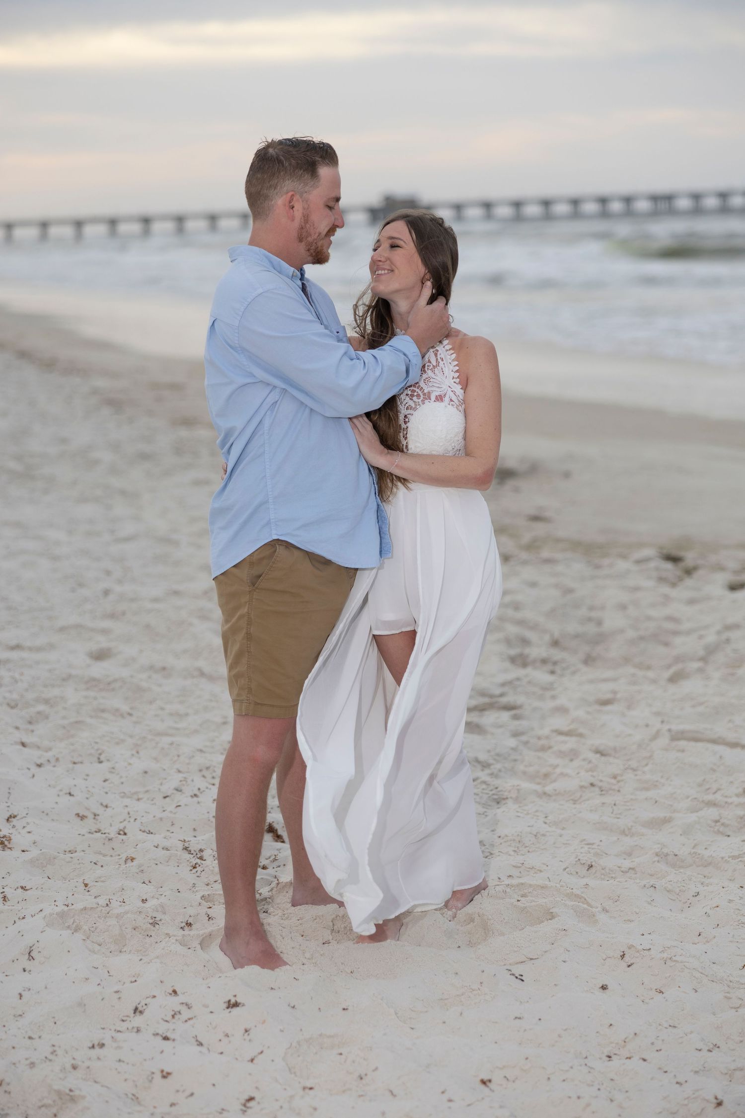 Couple embracing on a sandy beach at sunset, woman in white dress, man in blue shirt and tan shorts, pier in background.