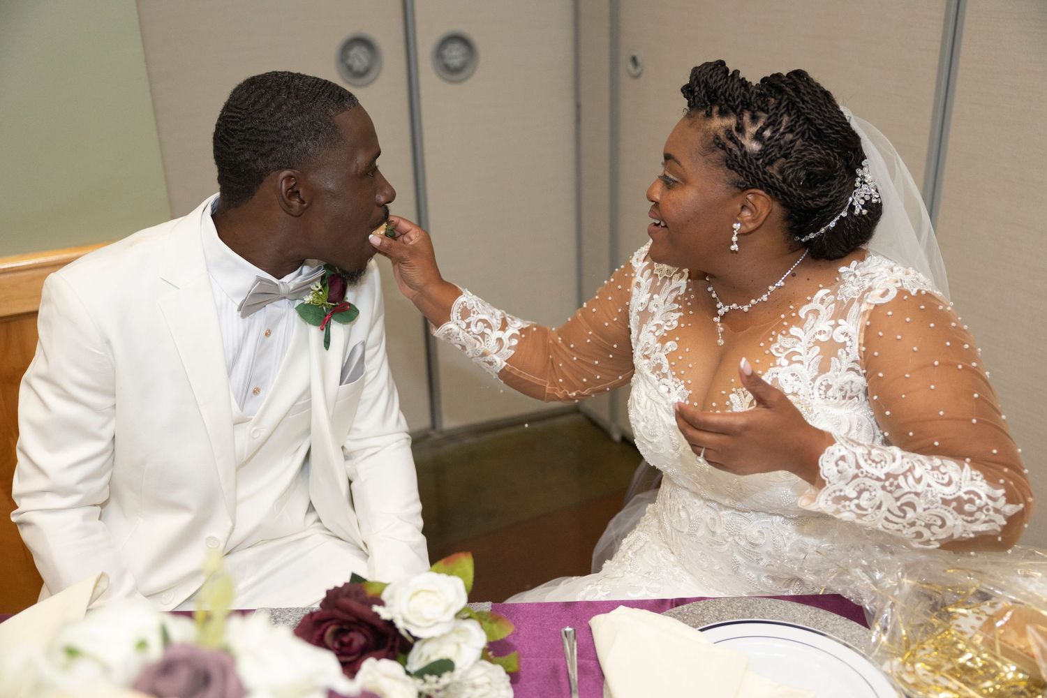 A bride in lace gown feeds cake to groom in white suit at wedding reception table with flowers.