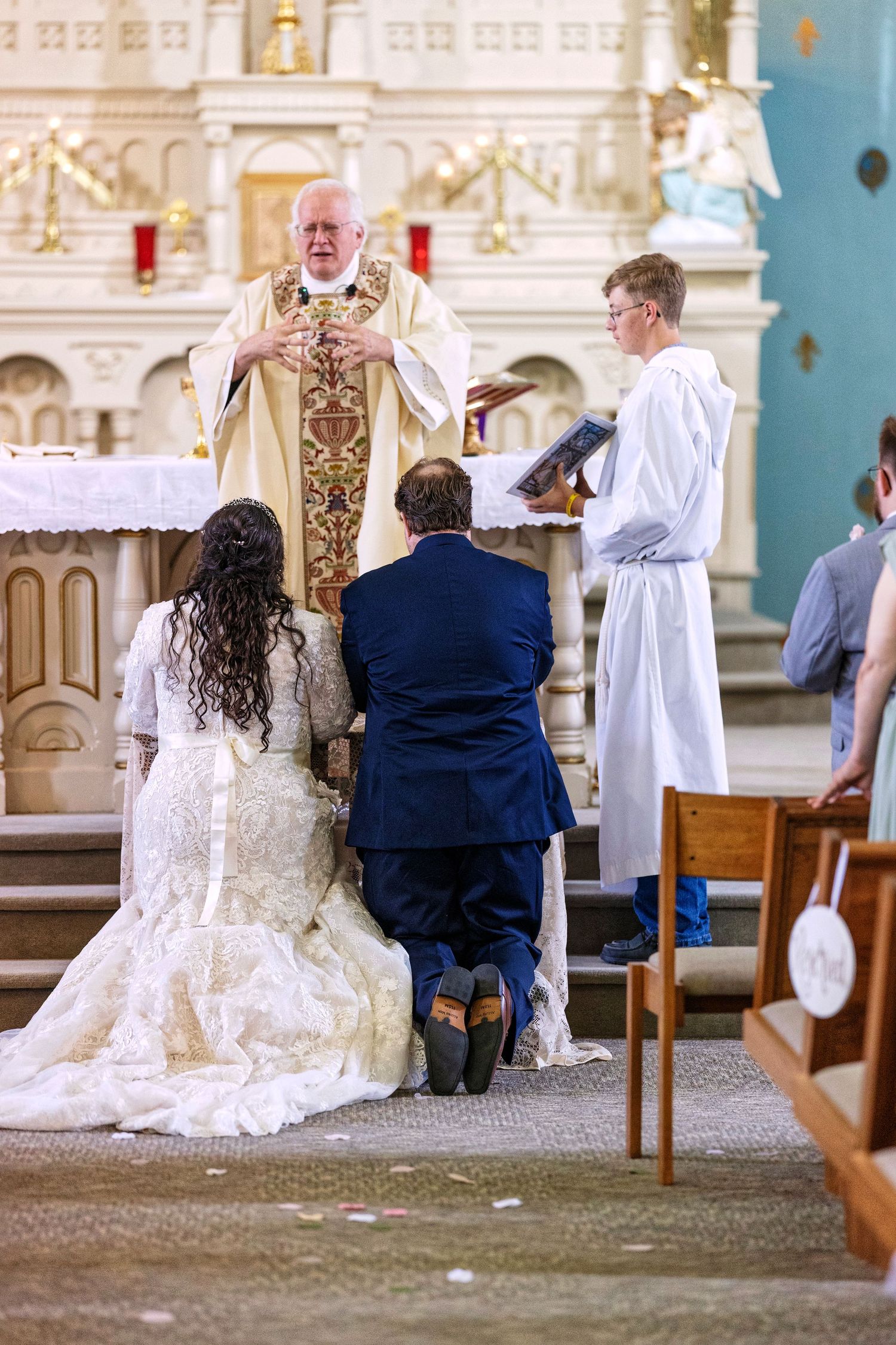 A wedding ceremony in progress inside an ornate church with couples kneeling at the altar in formal attire.