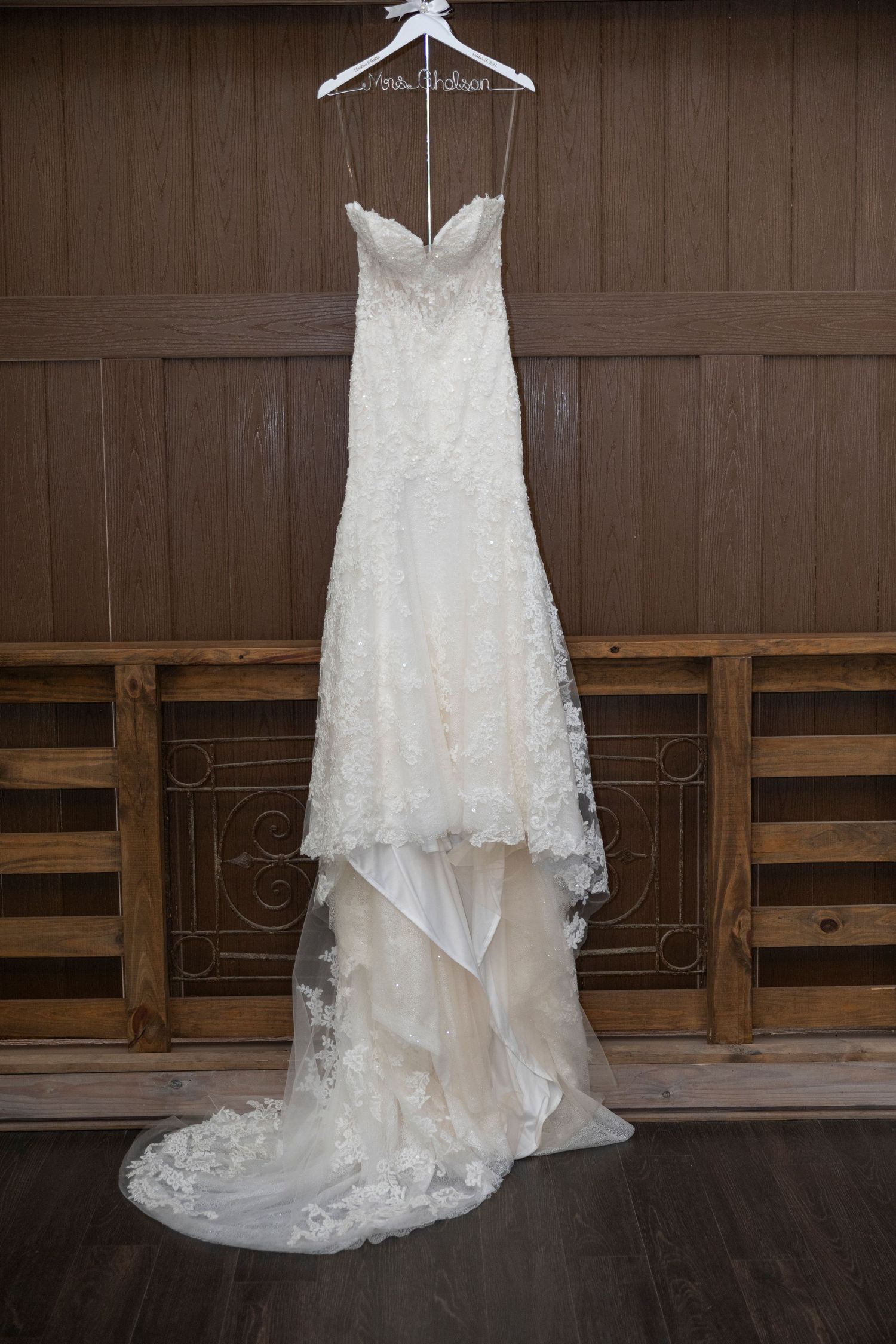 White lace wedding dress hanging on wooden wall with elegant train and delicate details displayed for photography.