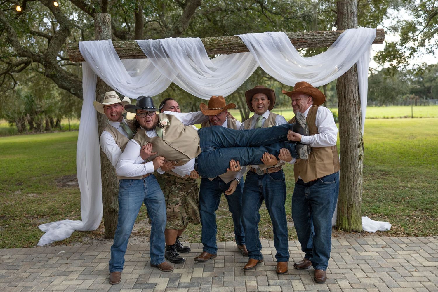 A group of western-styled performers pose together under a white draped arch at an outdoor event.