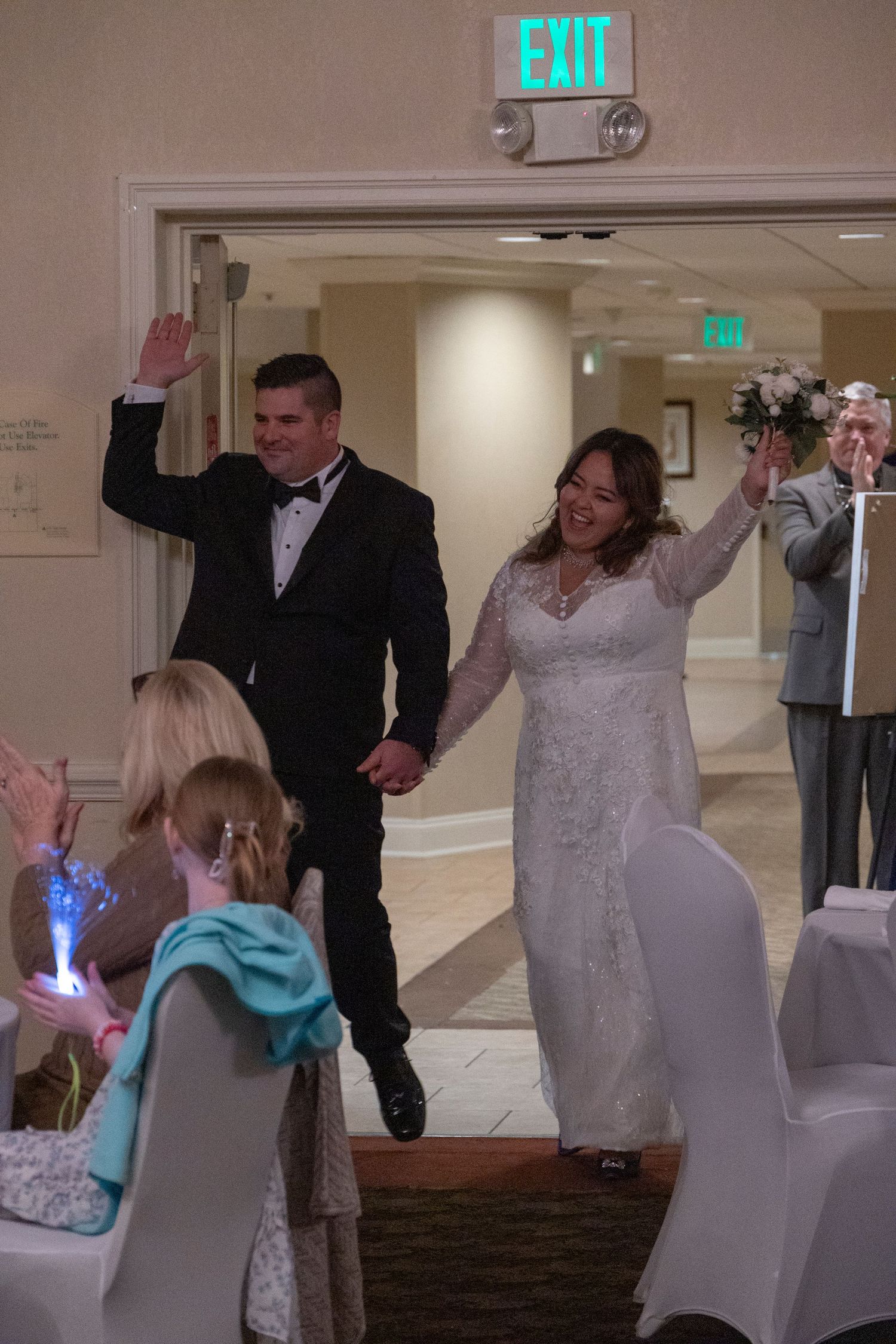 A couple makes their grand entrance at a wedding reception, walking through a hallway with an illuminated exit sign above.